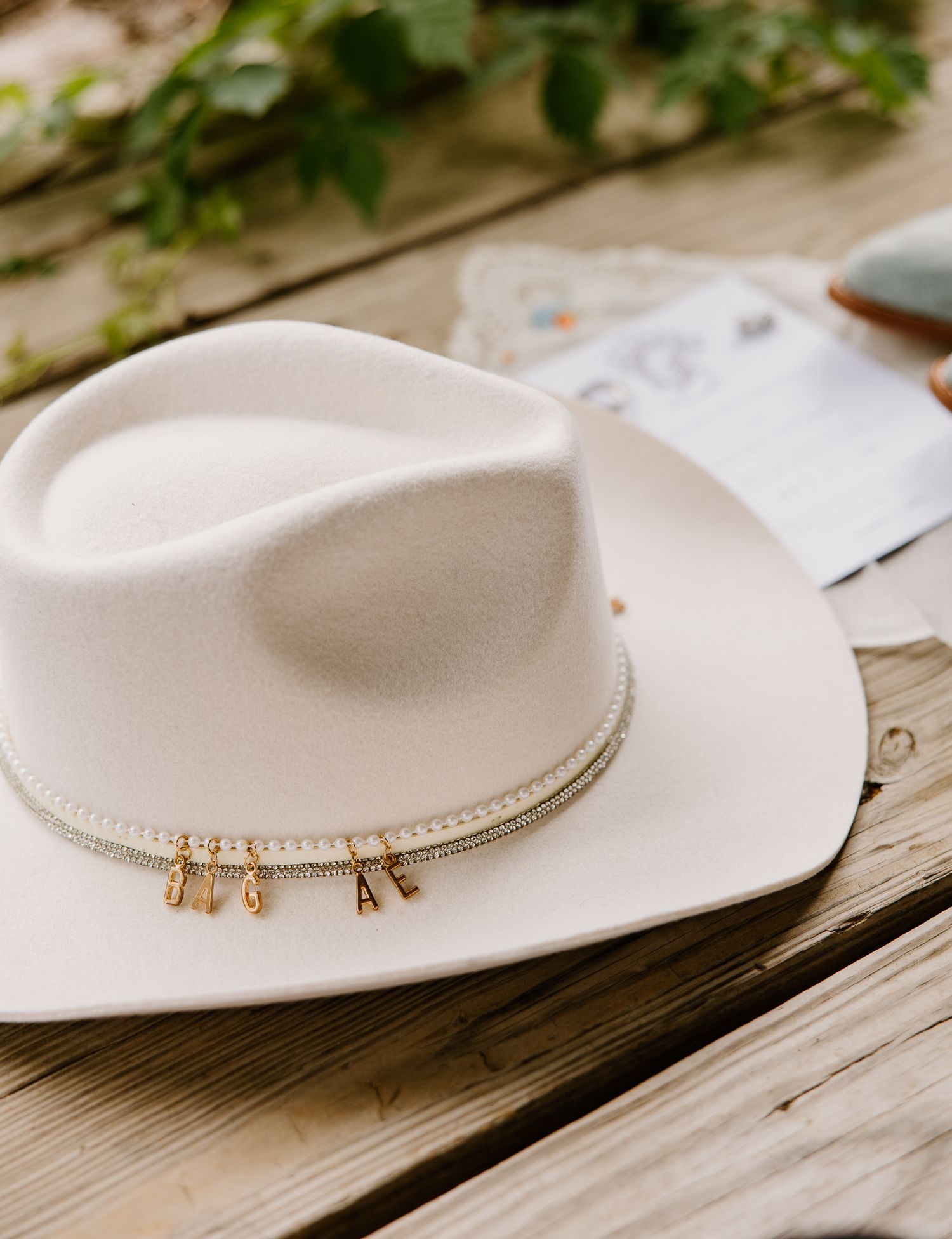 White felt cowboy hat with gold letter charms on braided band, displayed on rustic wooden surface.