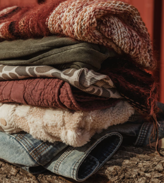 Neatly stacked pile of cozy fall clothing including knit sweaters, flannel, sherpa, and denim in warm tones.
