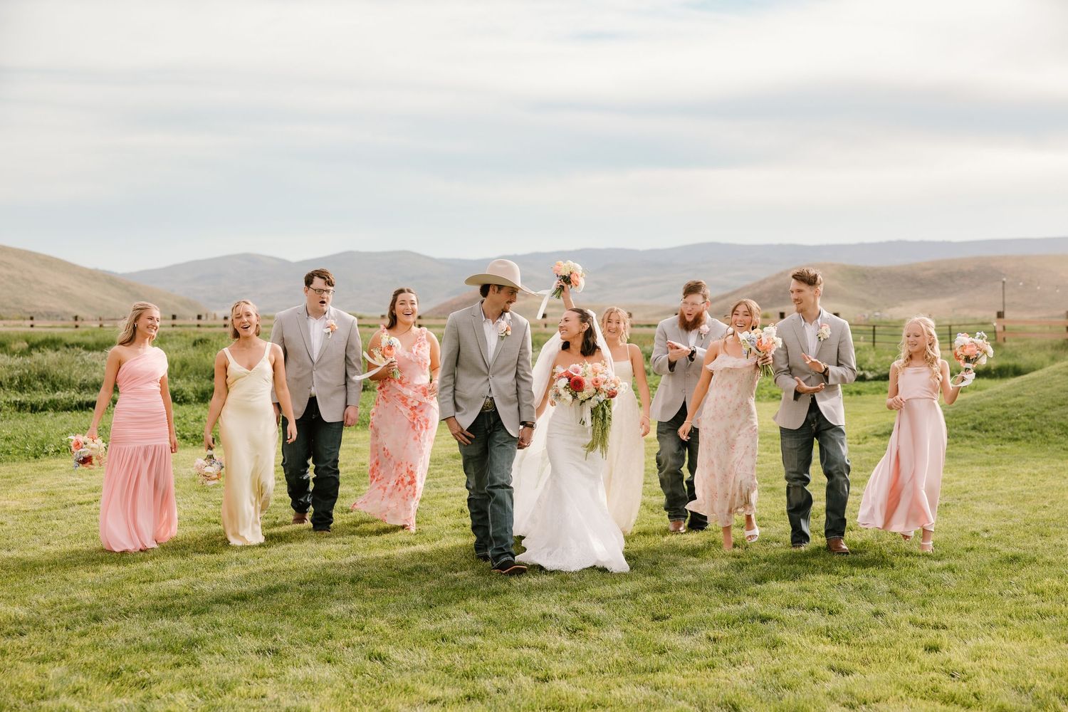Wedding party walking outdoors in green field with mountains, bridesmaids in pink dresses, groomsmen in gray suits.