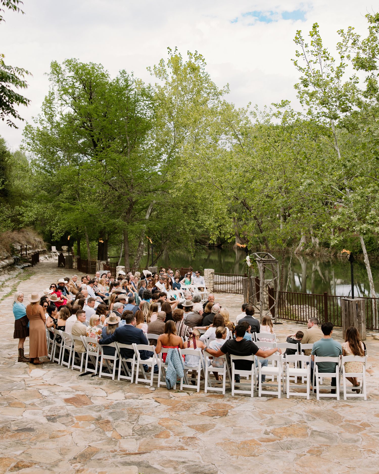 Outdoor wedding ceremony with guests seated on white chairs on a stone patio surrounded by lush green trees.