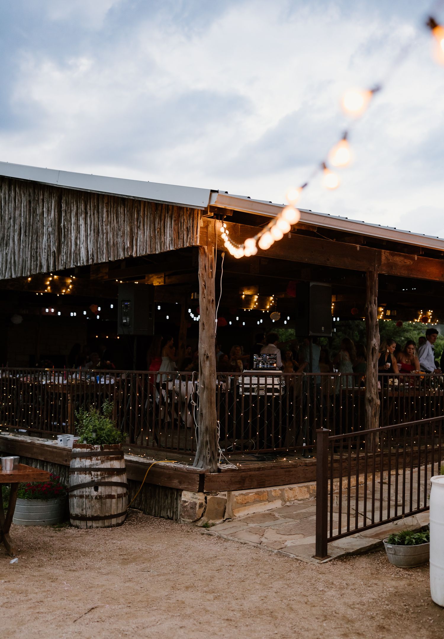 A rustic barn wedding venue glows with warm string lights at dusk, surrounded by wooden barrels and greenery.