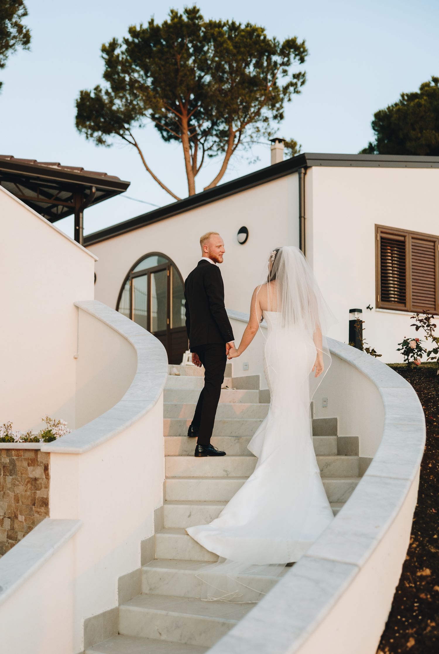 Bride in elegant mermaid gown with veil and groom in black tuxedo ascending curved white marble stairs.