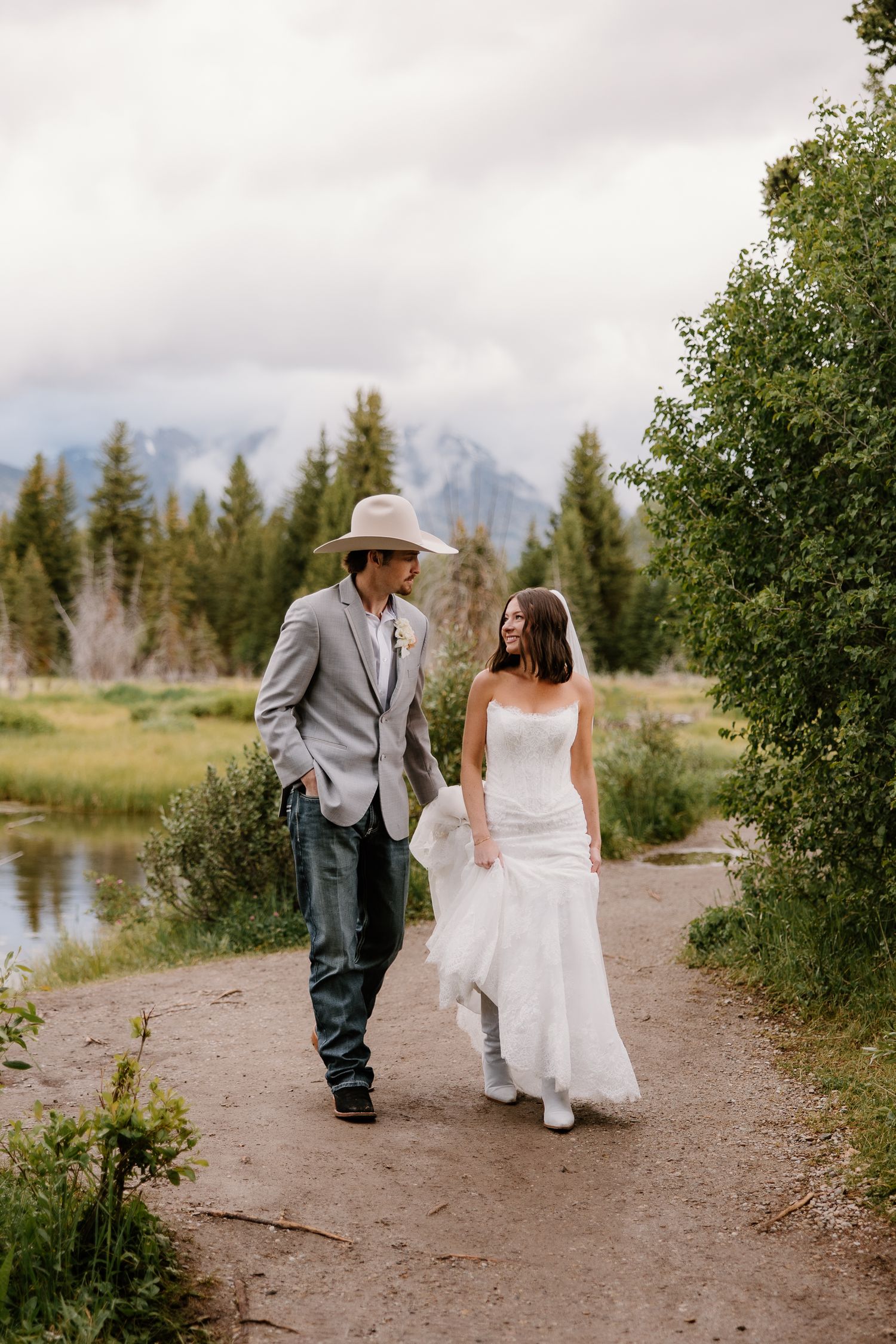 Bride in strapless white gown and groom in gray suit and cowboy hat walk together on scenic mountain trail.