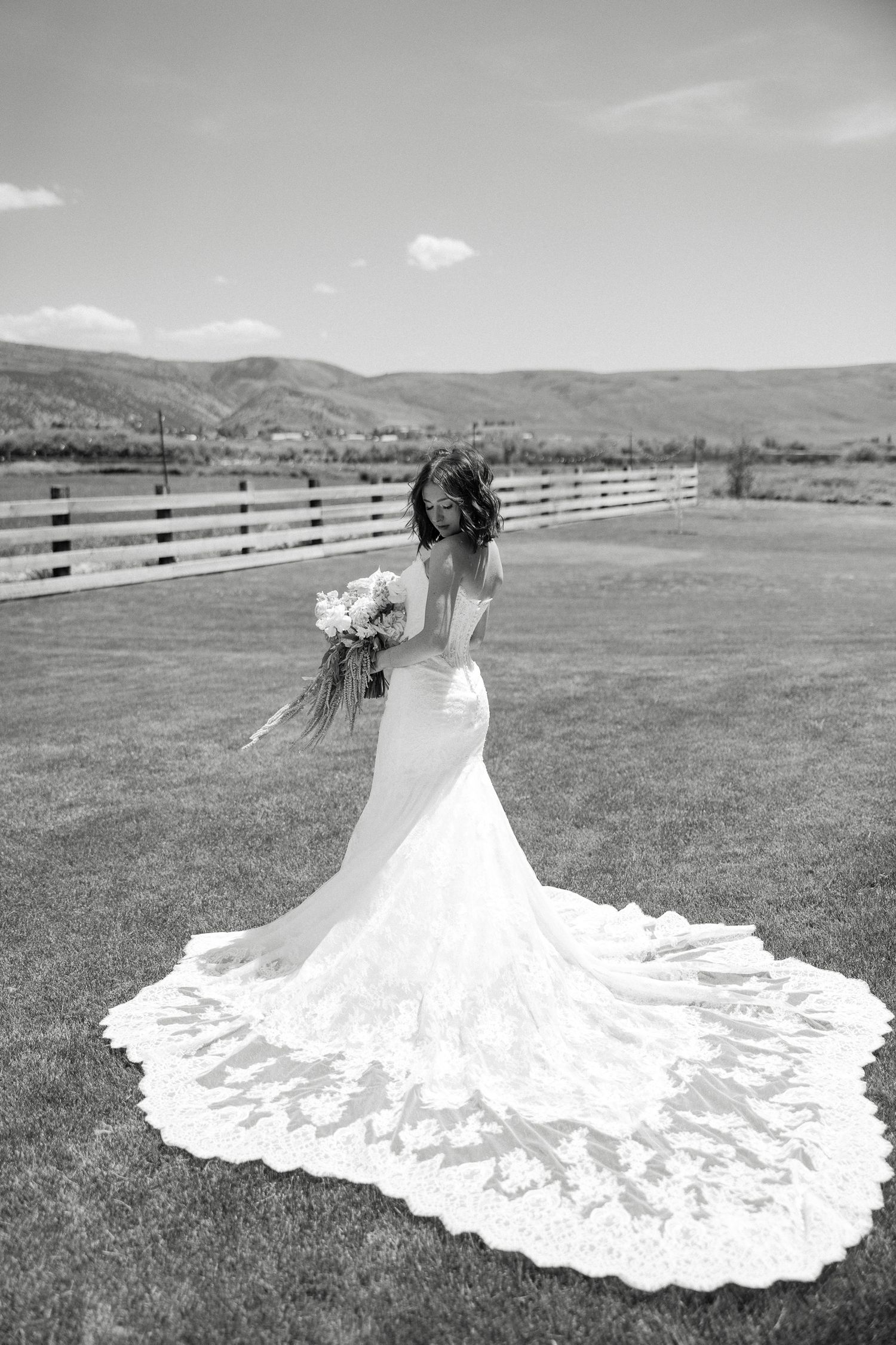 Black and white photo of a bride in a lace mermaid gown with long train holding bouquet in an open field.