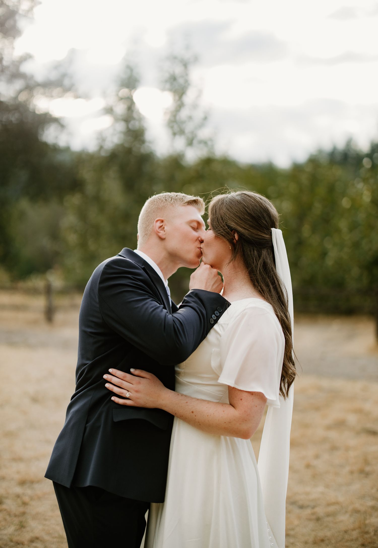 Groom in navy suit tenderly kisses bride in white dress in golden field with forest background.