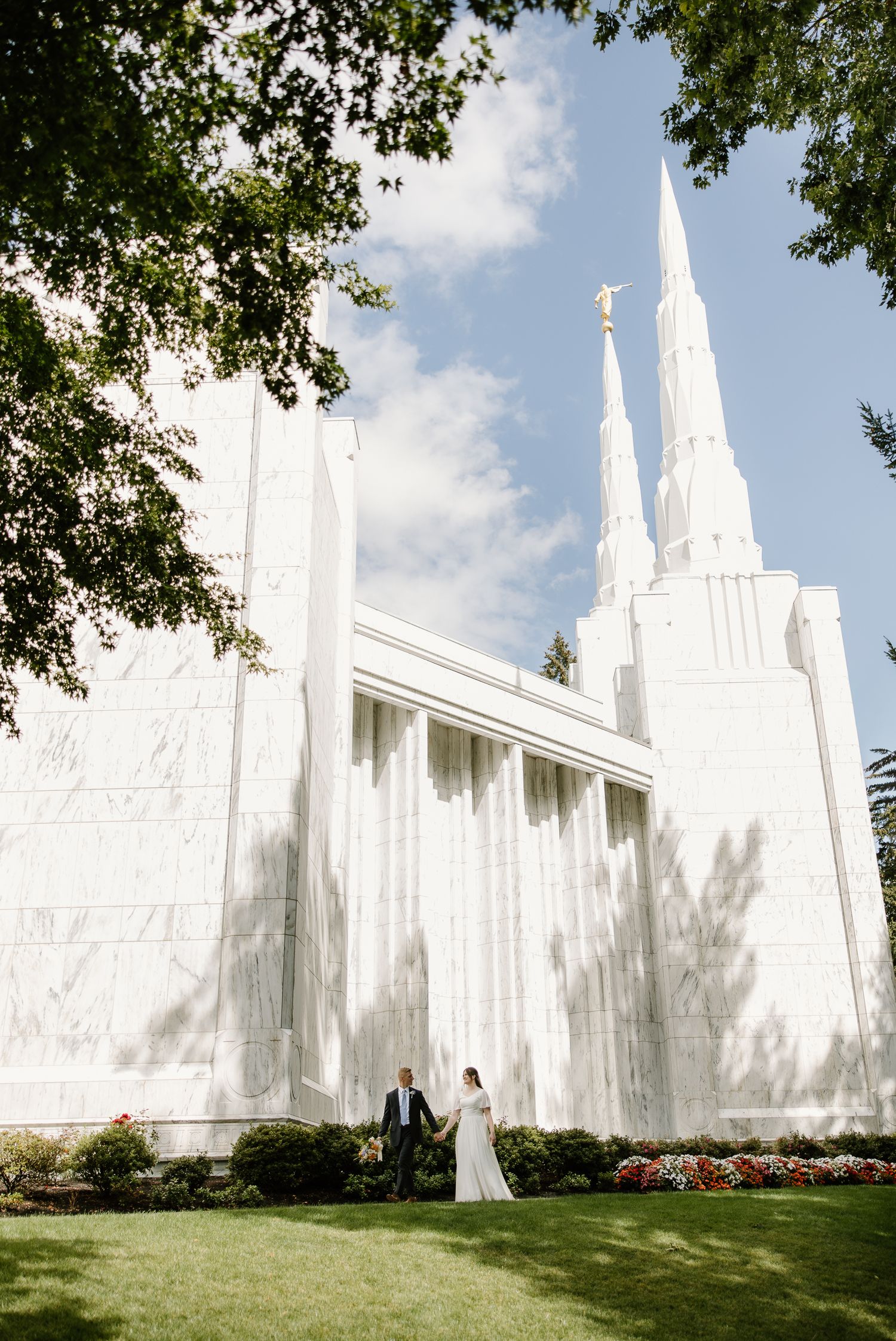 Bride and groom hold hands outside a white LDS temple with tall spires on a sunny day.
