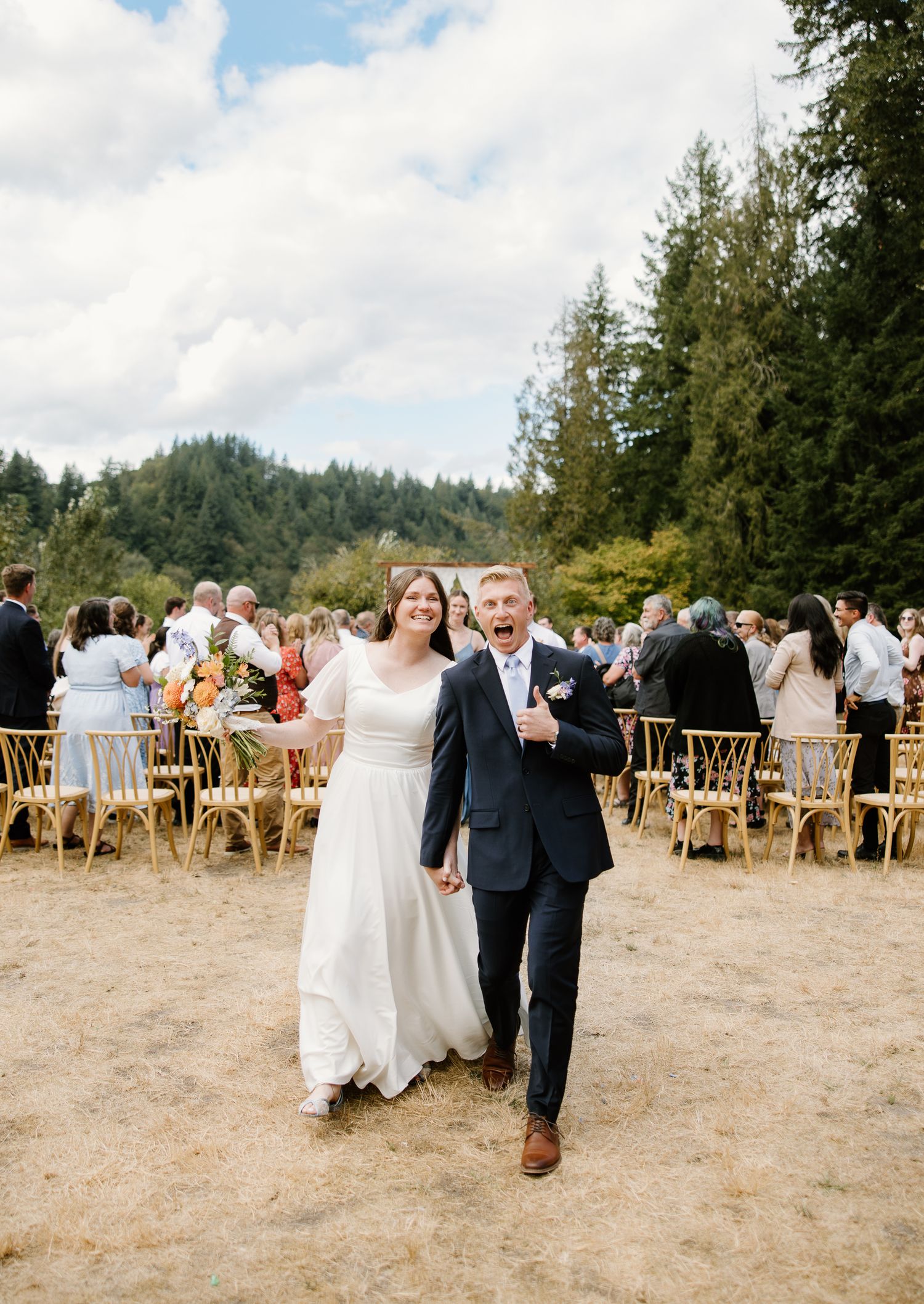 Joyful newlyweds walk back down the aisle after outdoor wedding ceremony with forest backdrop.