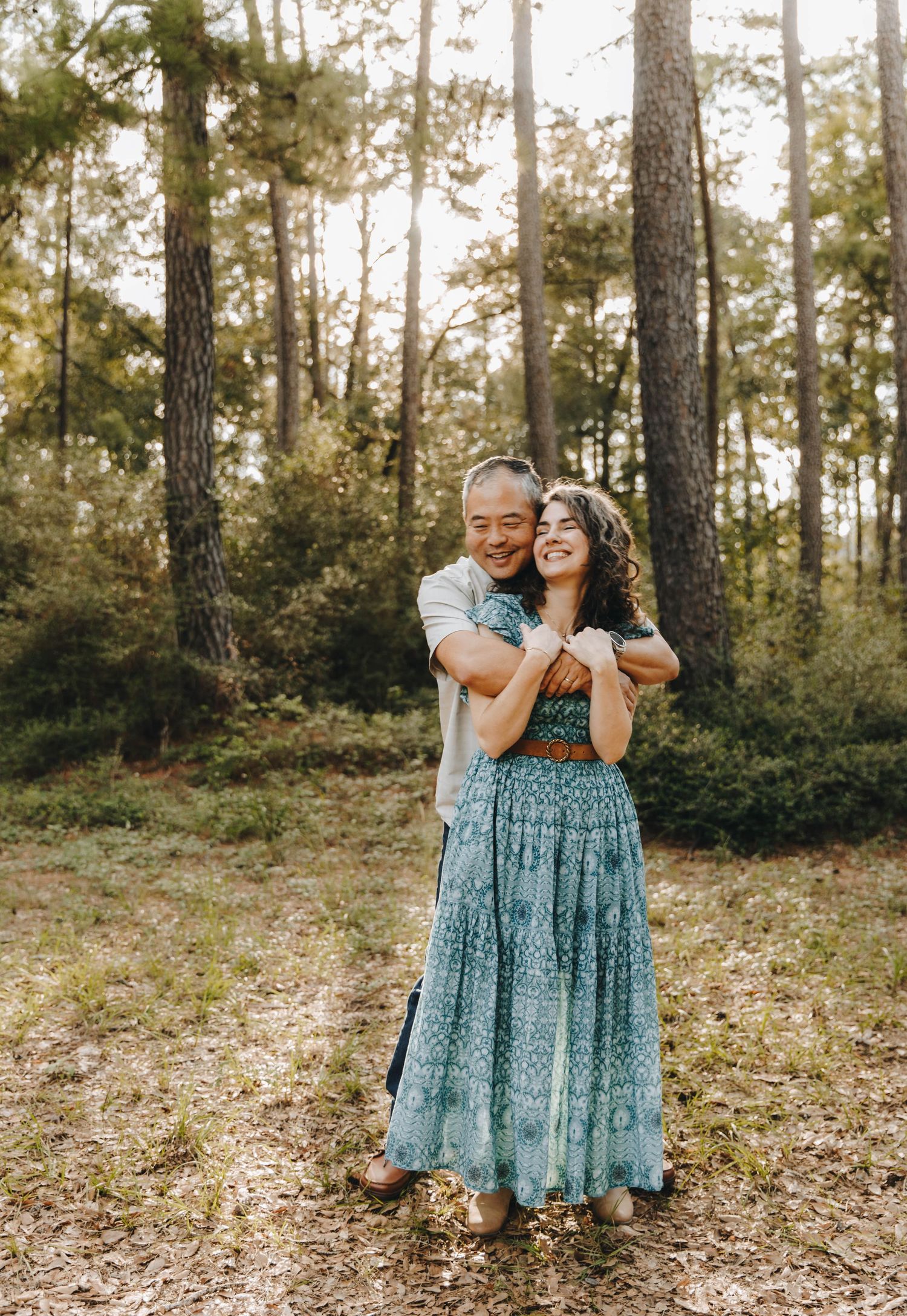 Couple embracing in a forest, woman in teal bohemian maxi dress, man hugging her from behind.