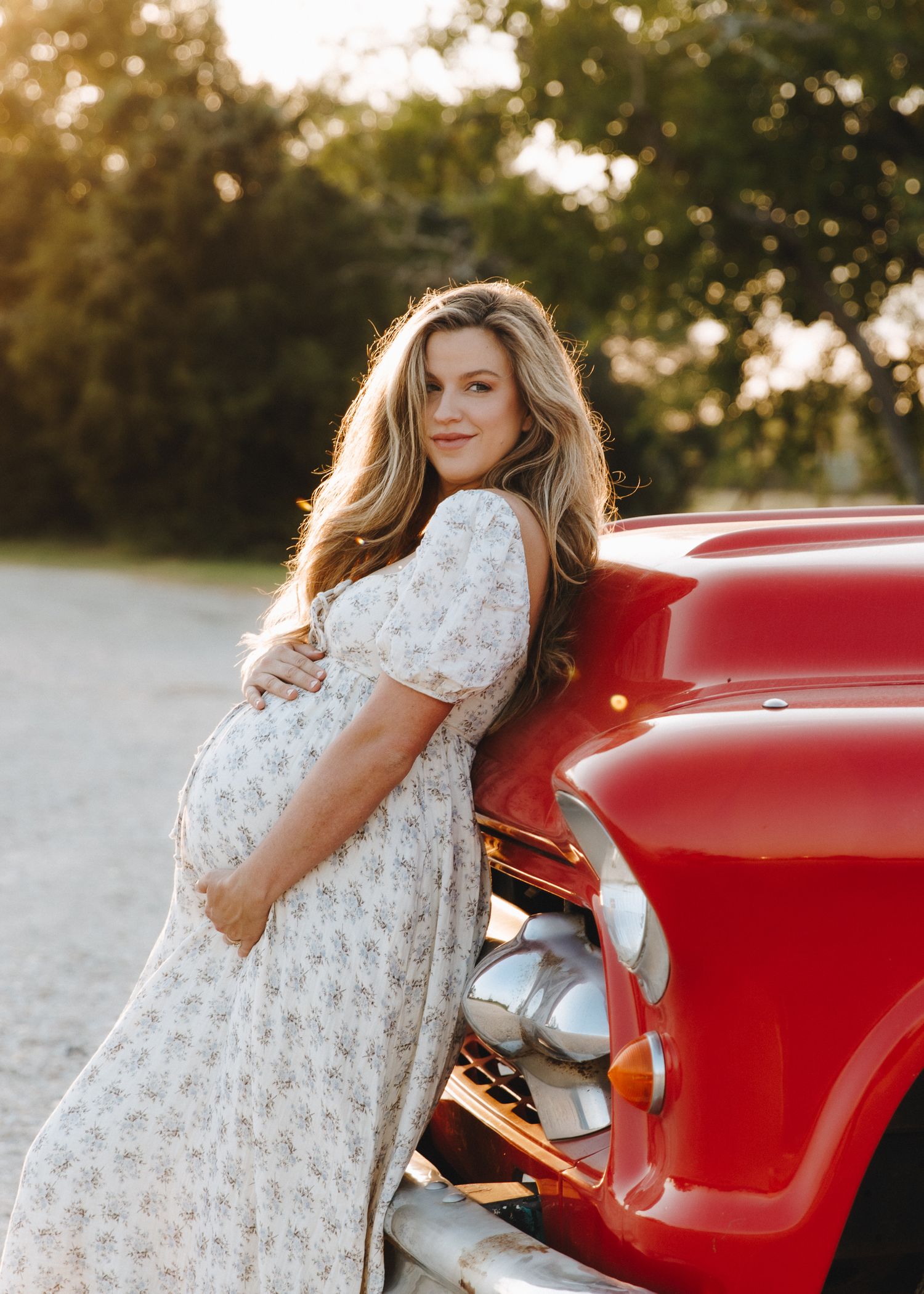 Pregnant woman in floral dress leaning against red vintage truck at golden hour.