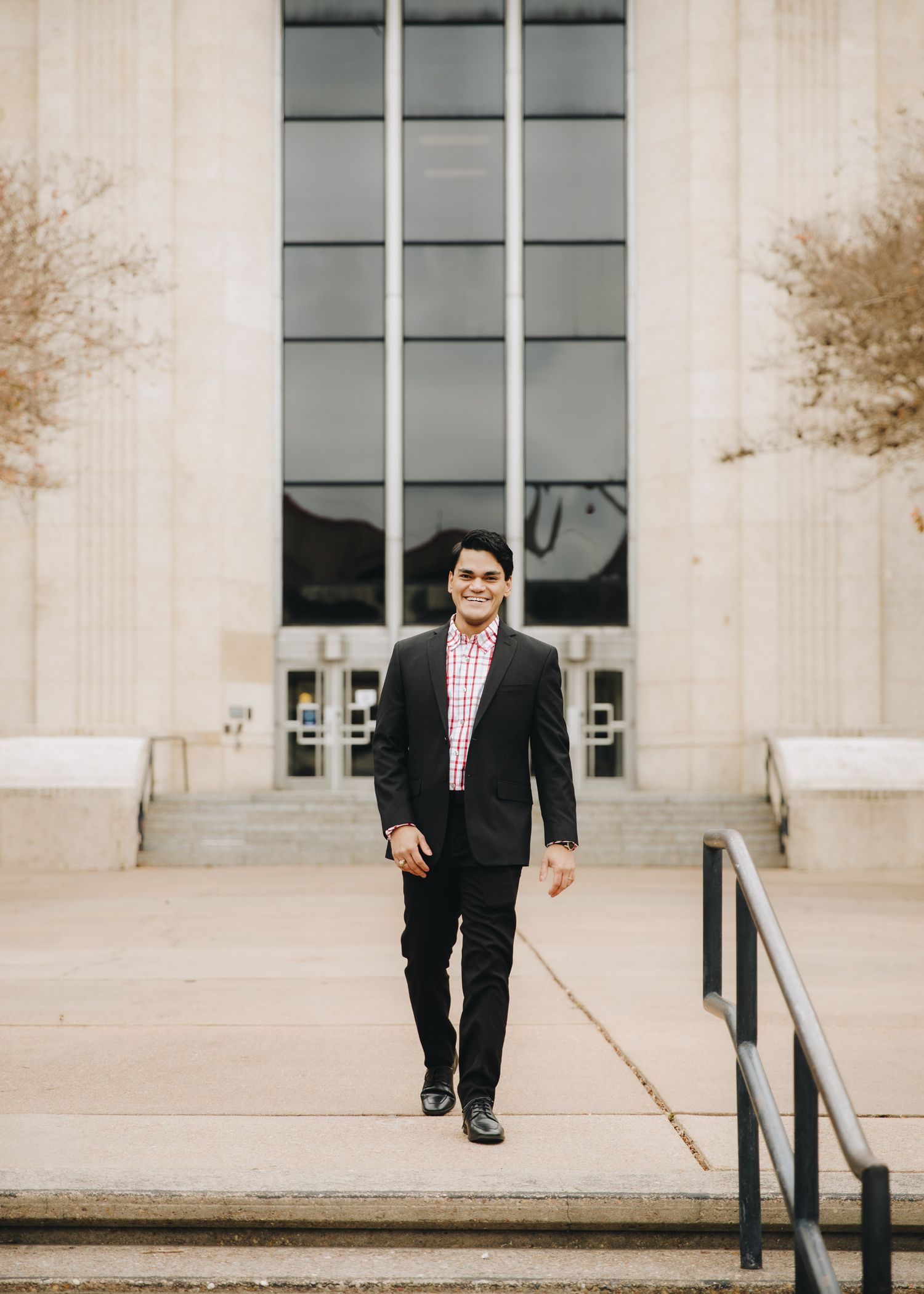 Smiling young man in black suit and plaid shirt walking down steps of large building.