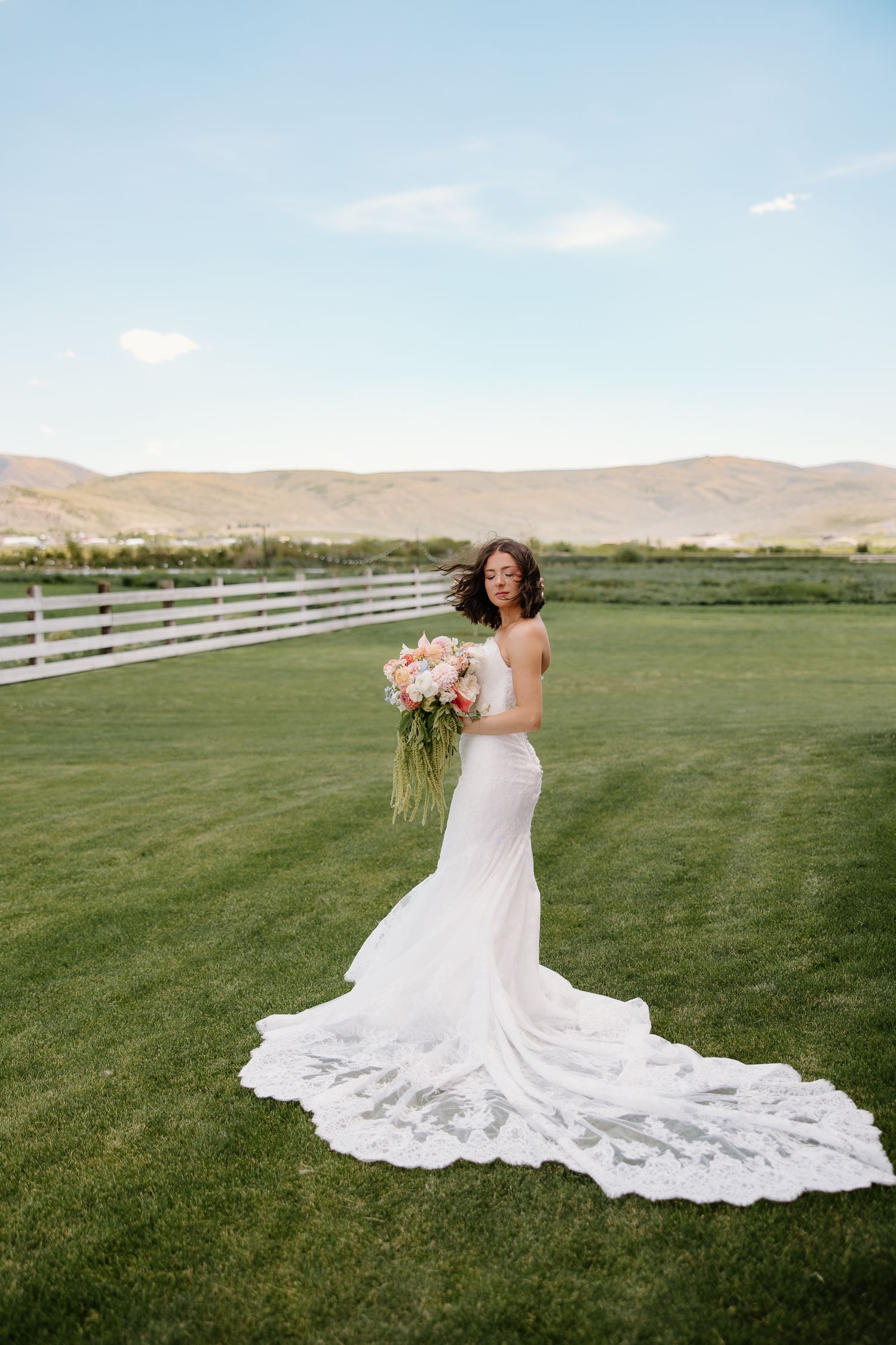 Bride in fitted lace gown with long train holding pink bouquet on green farm lawn.