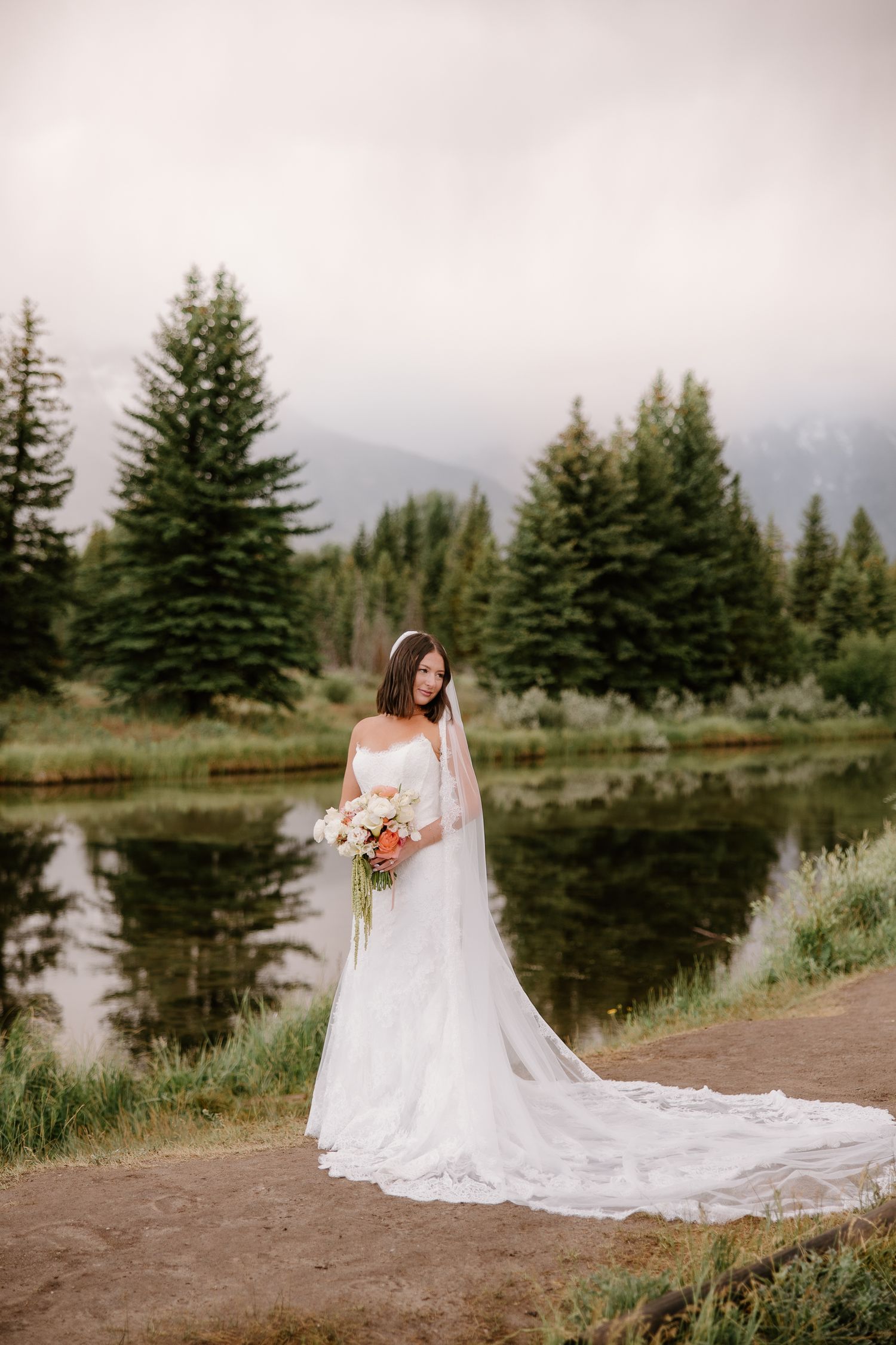 Bride in fitted lace gown with long veil holding bouquet beside reflective mountain pond.