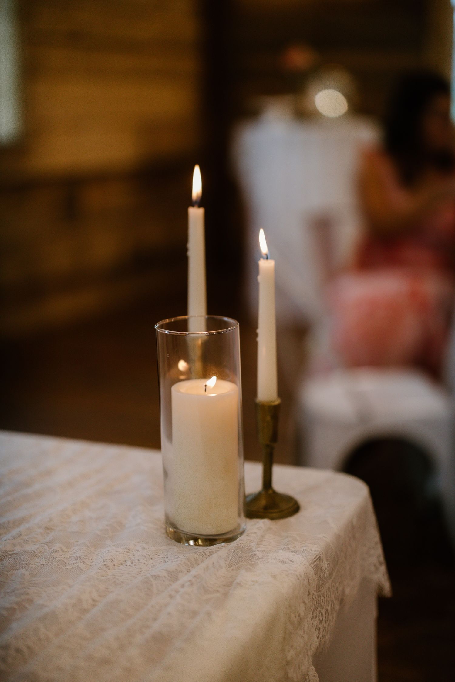 Unity candle ceremony setup with lit white pillar and taper candles on lace-covered table indoors.
