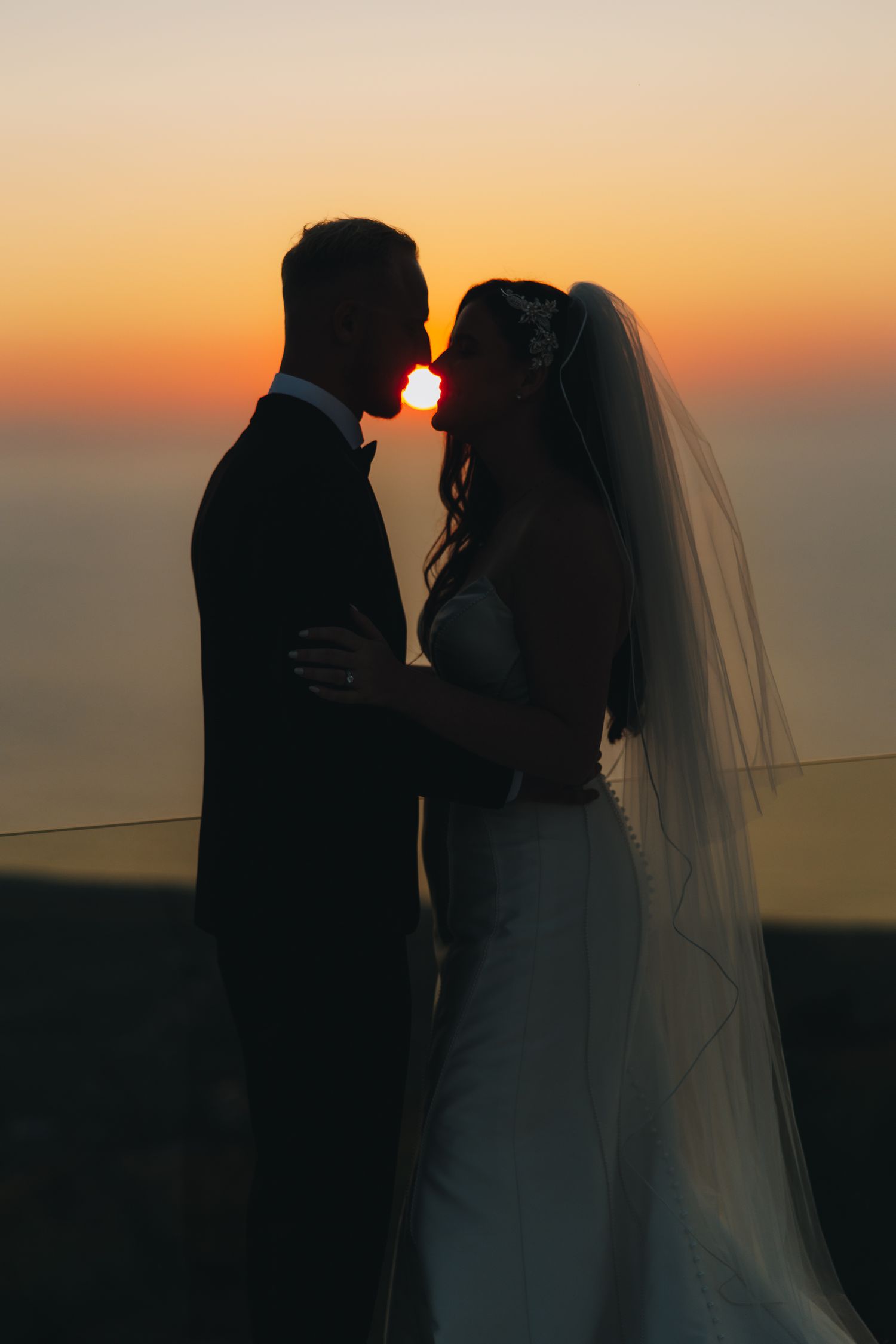 Silhouette of bride and groom sharing a romantic moment against a vibrant orange sunset over the ocean.