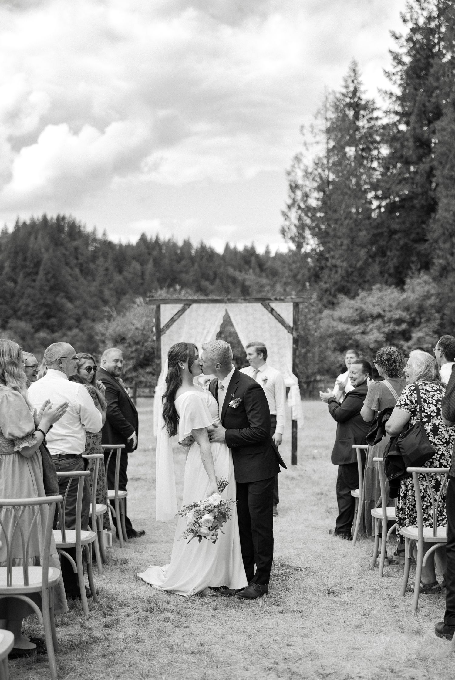 Black and white photo of bride and groom sharing first kiss at outdoor wedding ceremony.