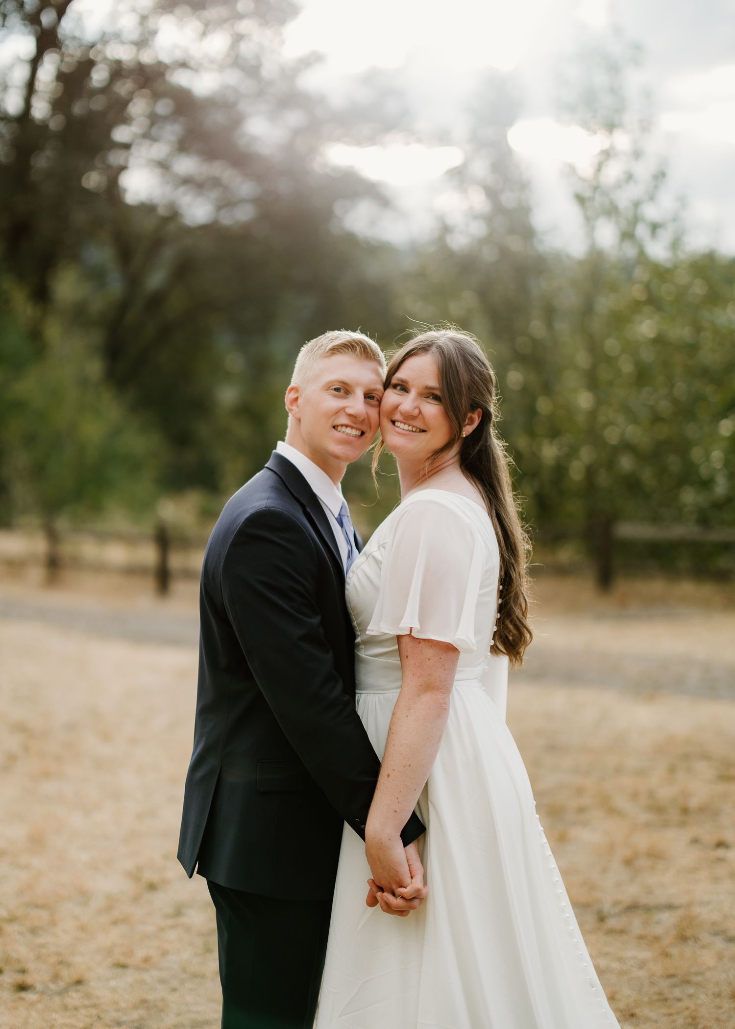 Smiling newlywed couple holds hands in golden orchard light, bride in flutter-sleeve white gown.