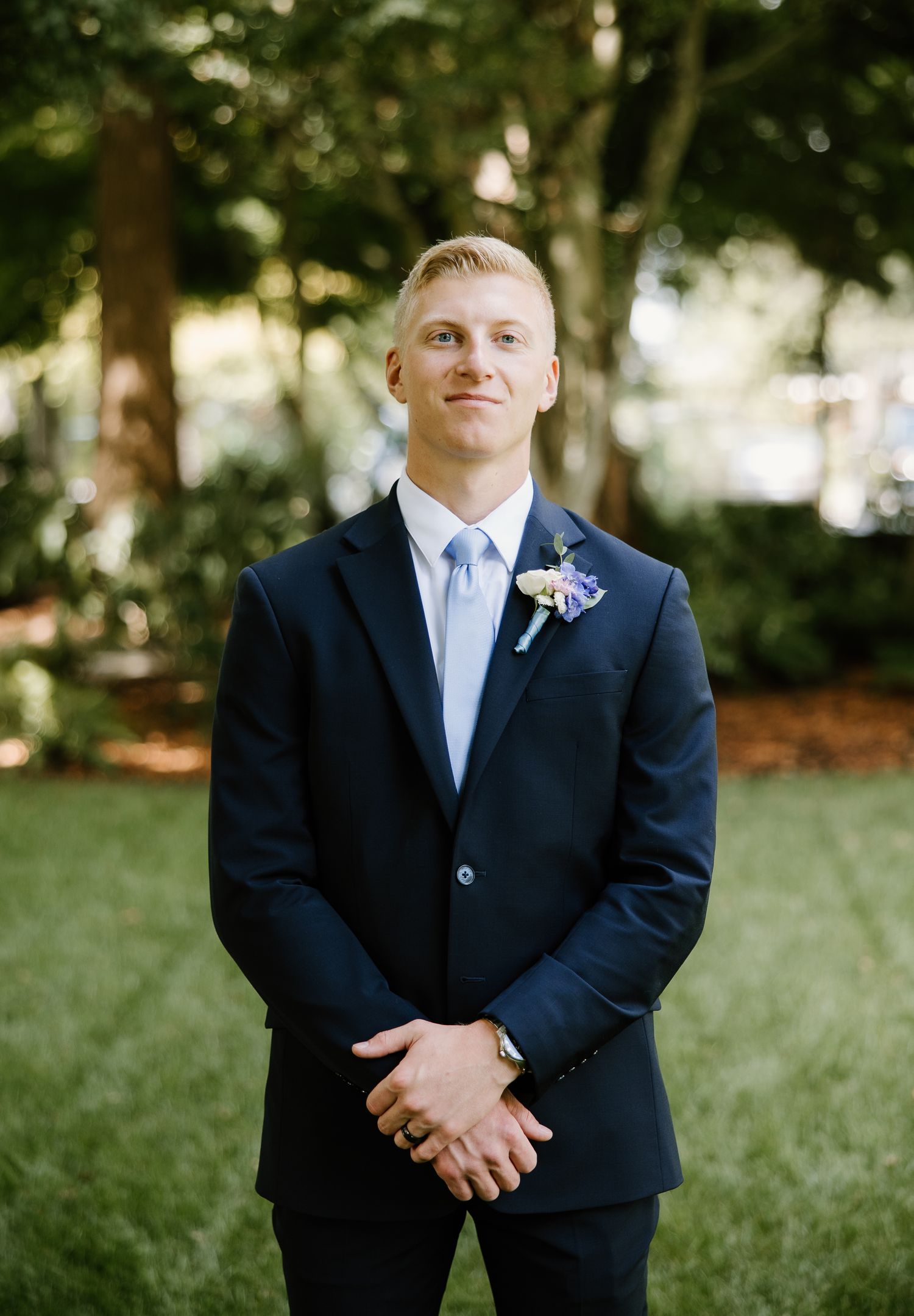 Groom in navy suit with boutonniere smiles outdoors on his wedding day.
