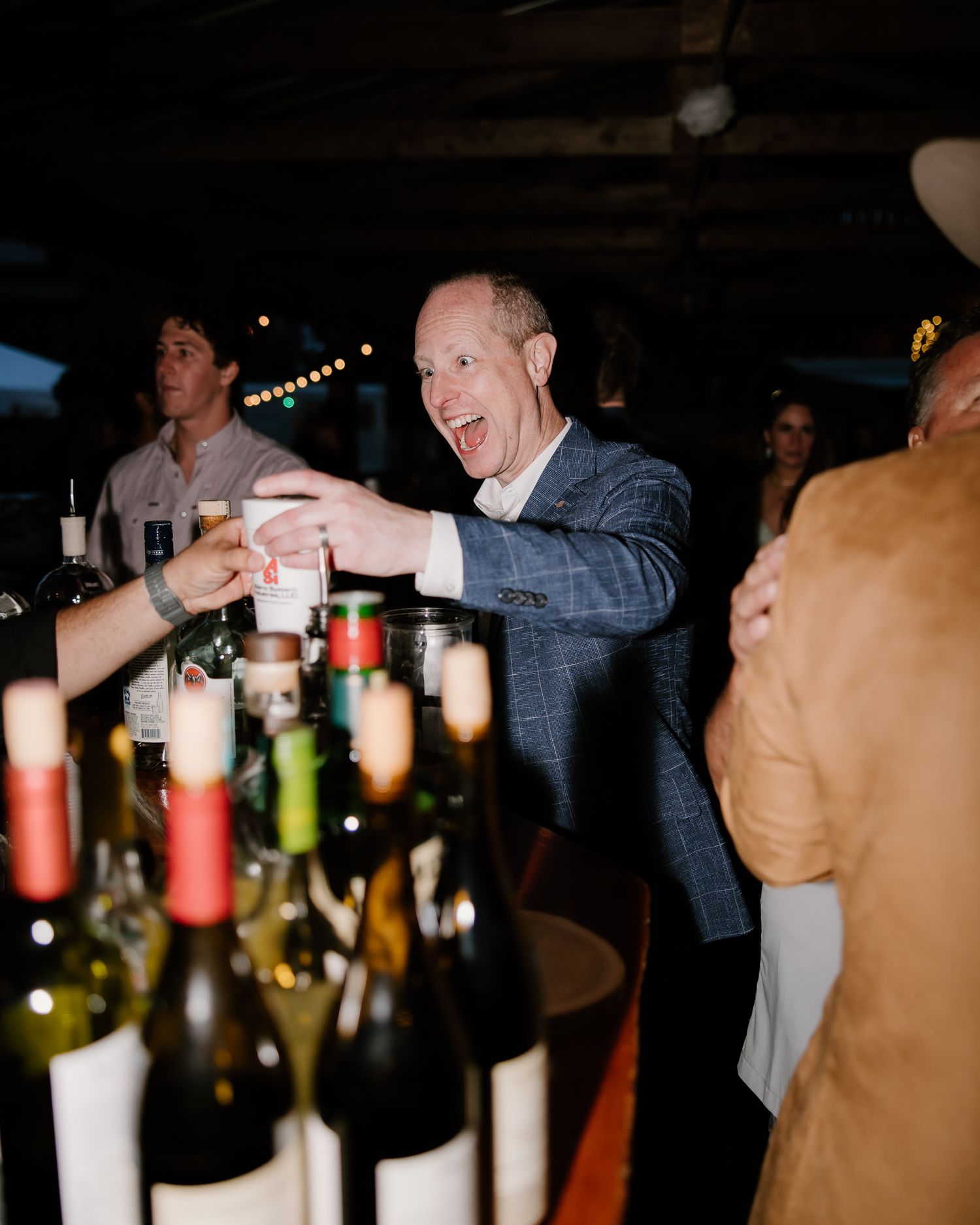 Smiling wedding guest in blue suit raises a toast at dimly lit reception bar surrounded by wine bottles.