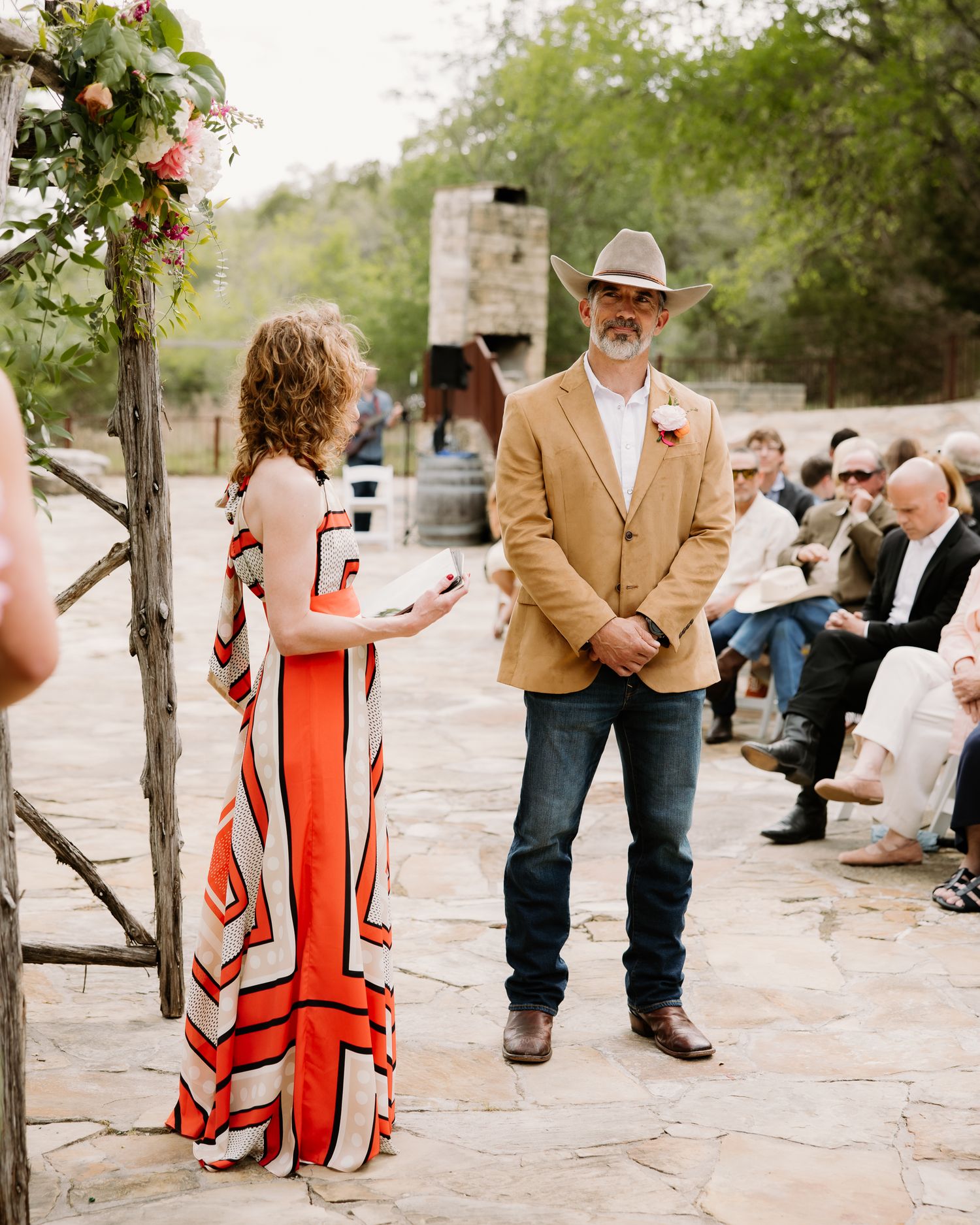 A wedding officiant in a colorful striped dress reads vows as the groom in a tan blazer and cowboy hat listens.