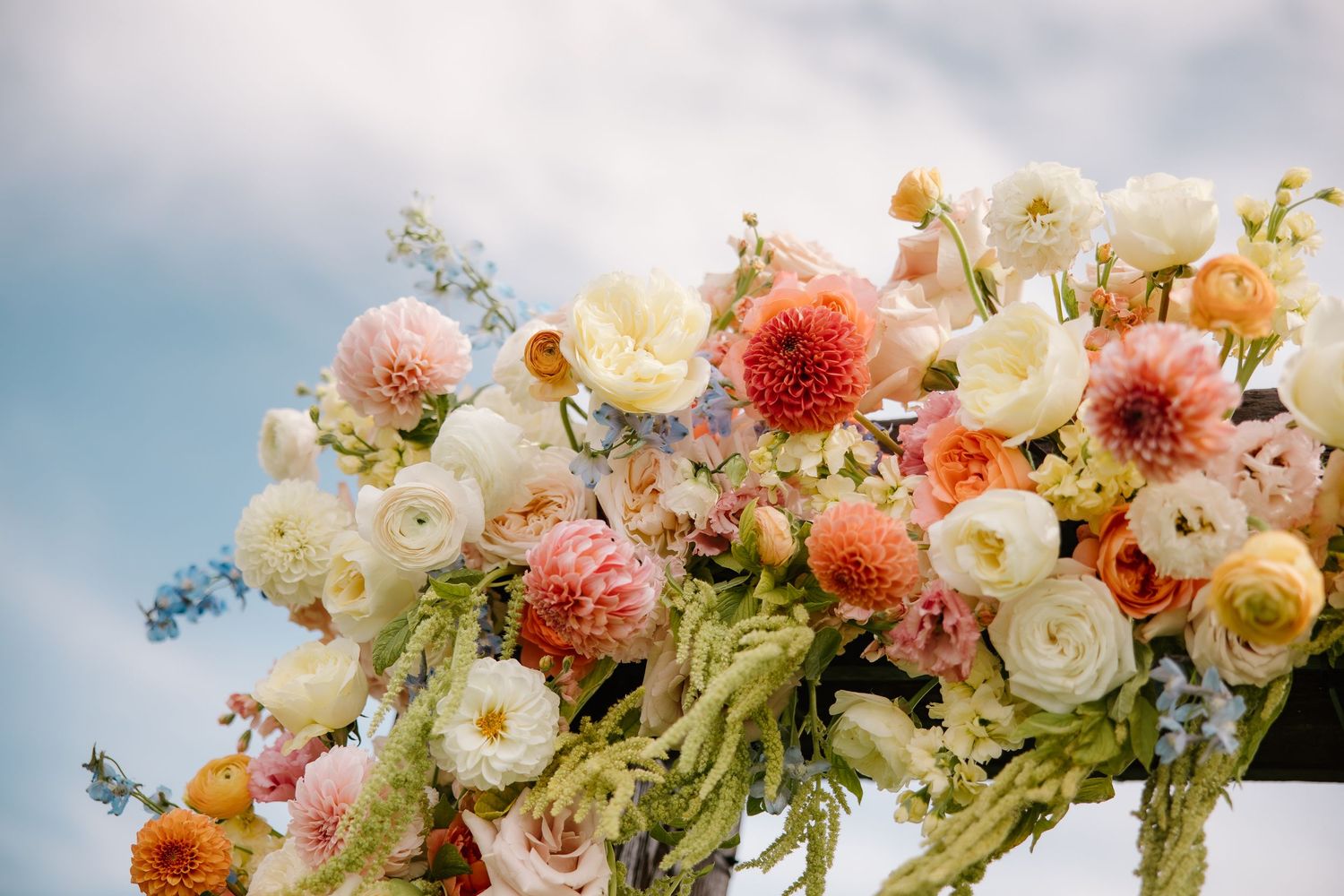 Colorful floral wedding arch arrangement with dahlias, ranunculus, and roses against a blue sky.