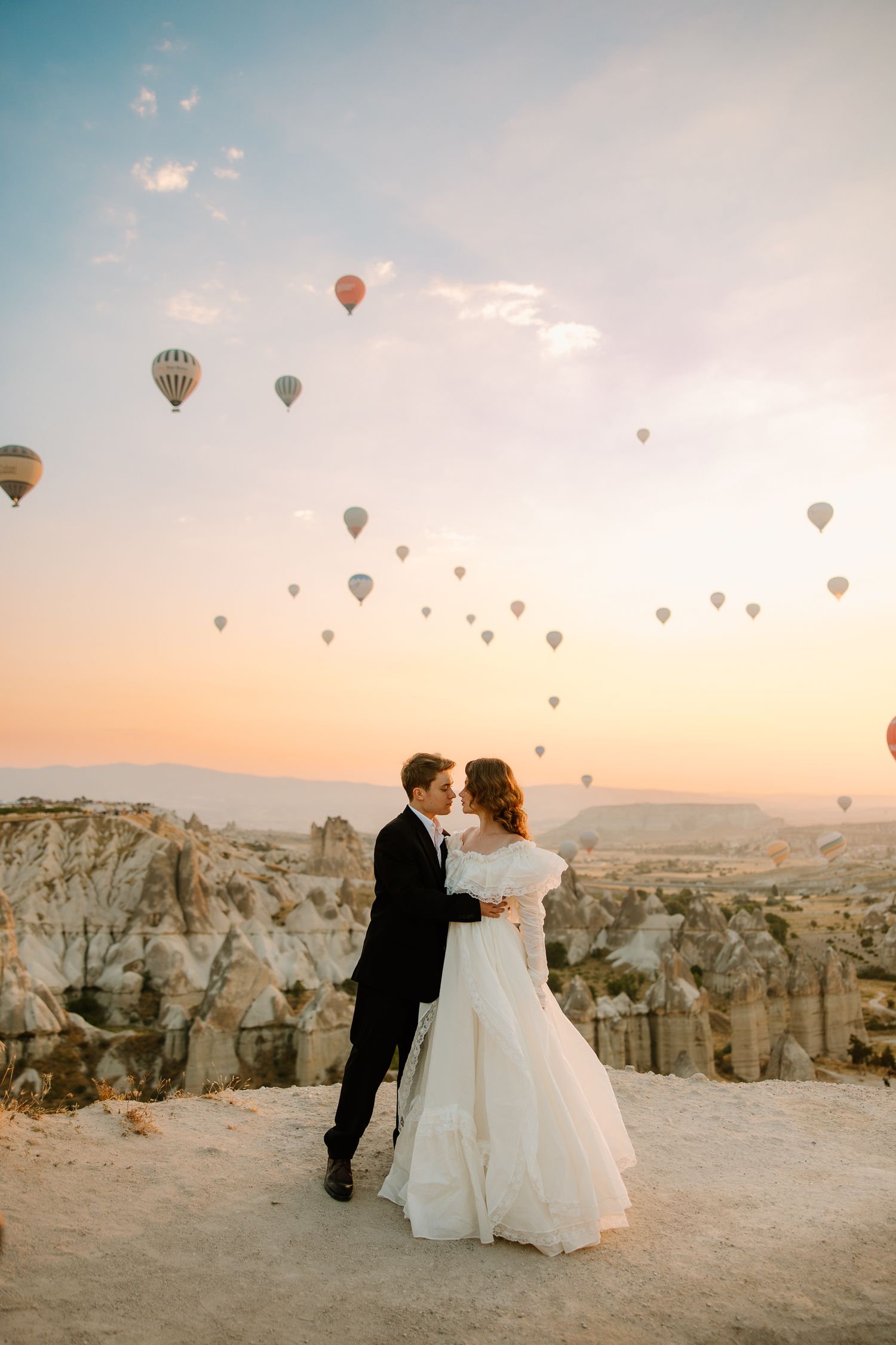 Bride and groom embrace at sunrise in Cappadocia with colorful hot air balloons floating over rocky landscape.
