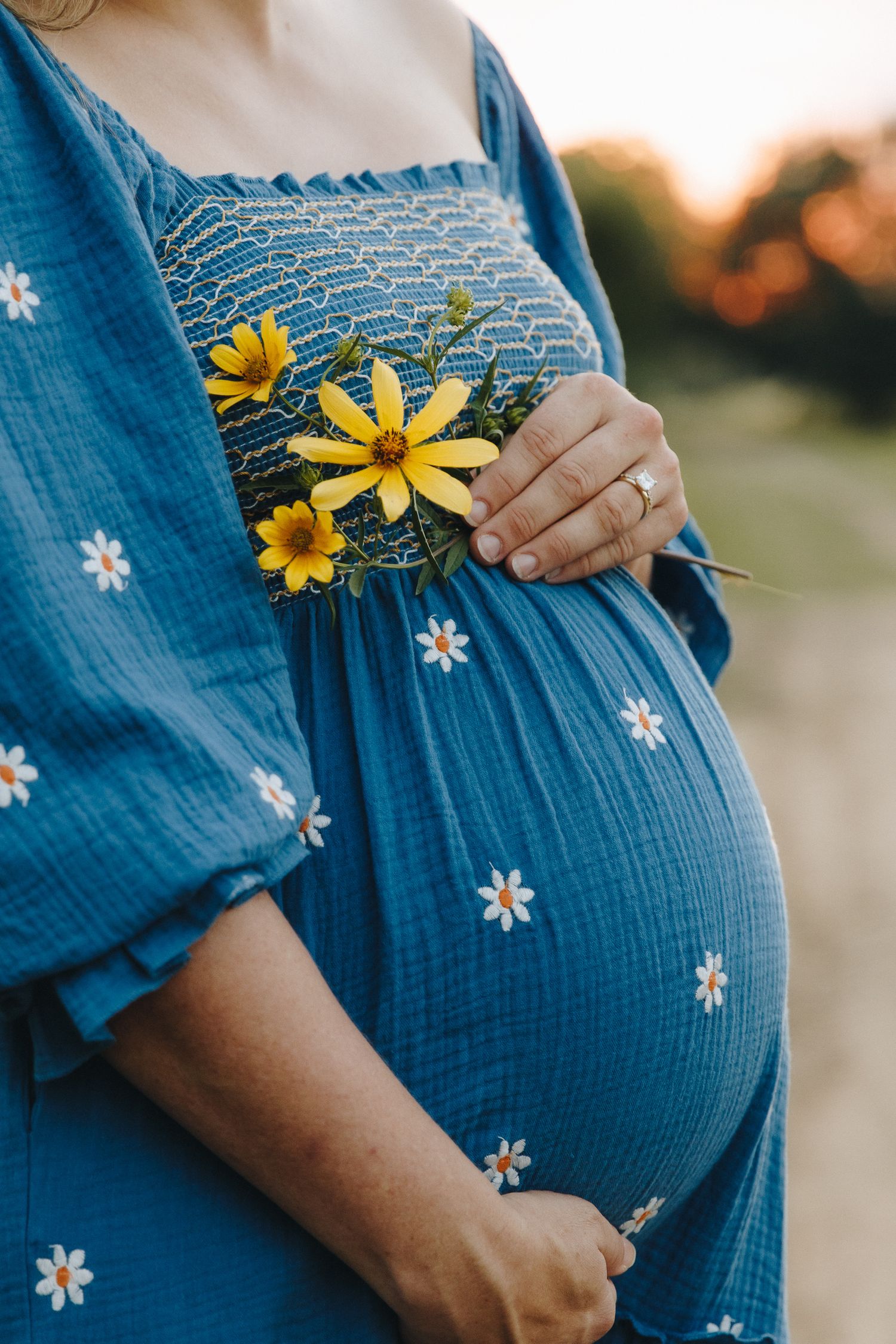 Close-up of pregnant belly in blue daisy dress with yellow wildflowers at golden hour.
