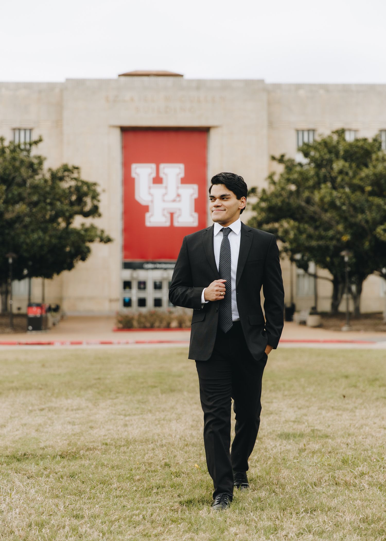 Young man in black suit smiling on campus lawn in front of University of Houston sign.