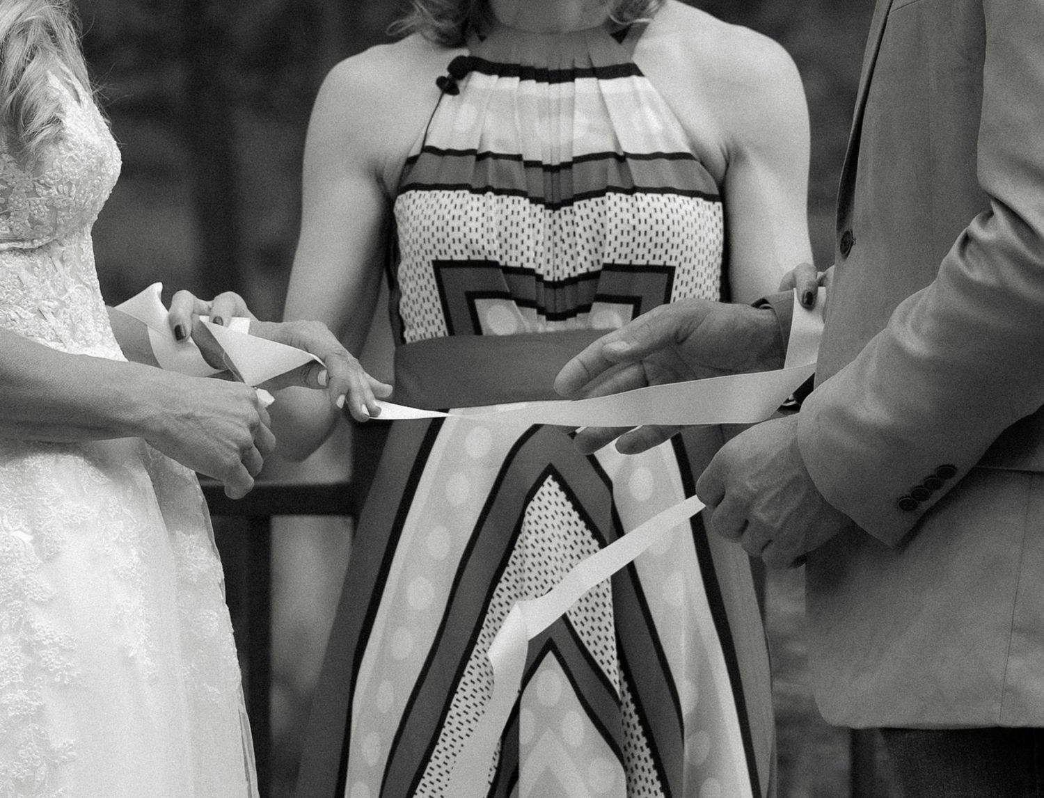 Black and white photo of wedding officiant in patterned dress performing ring exchange ceremony between bride and groom.