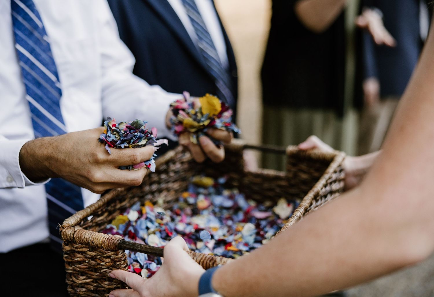 Guests grabbing colorful confetti from a wicker basket at a wedding celebration outdoors.