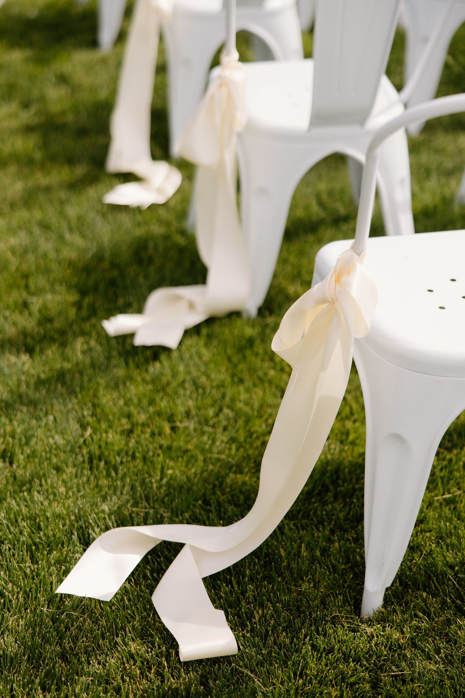 White chairs with ivory ribbon bows on green grass at an outdoor wedding ceremony.