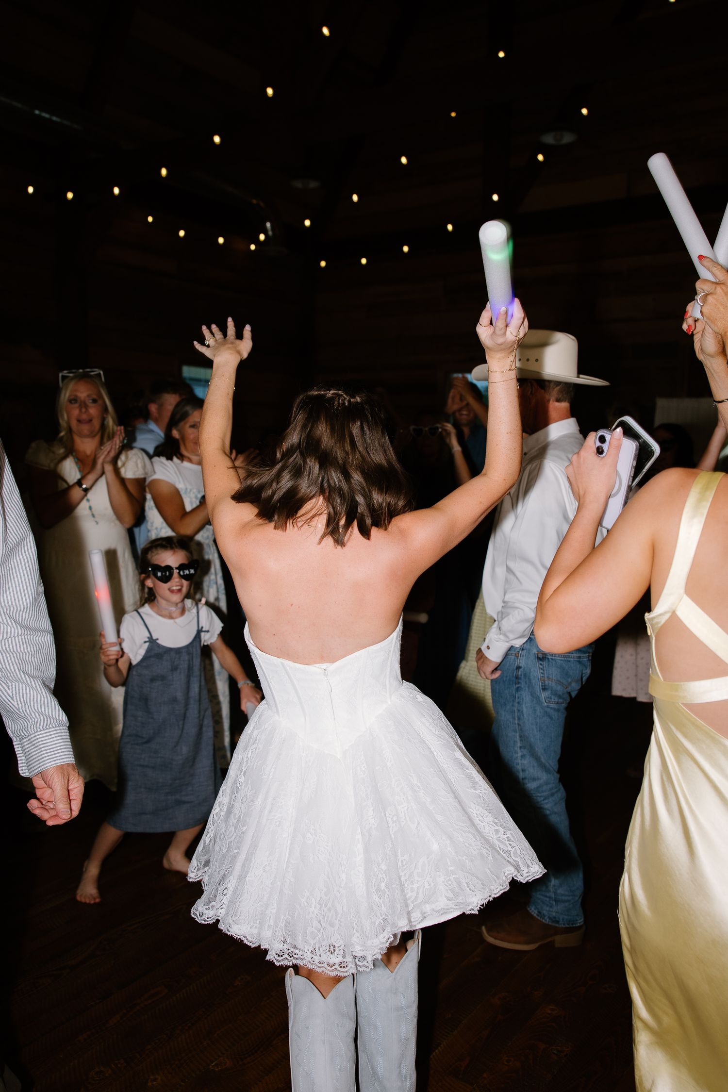 Bride in short white dress dancing with glow sticks raised at lively wedding reception party.