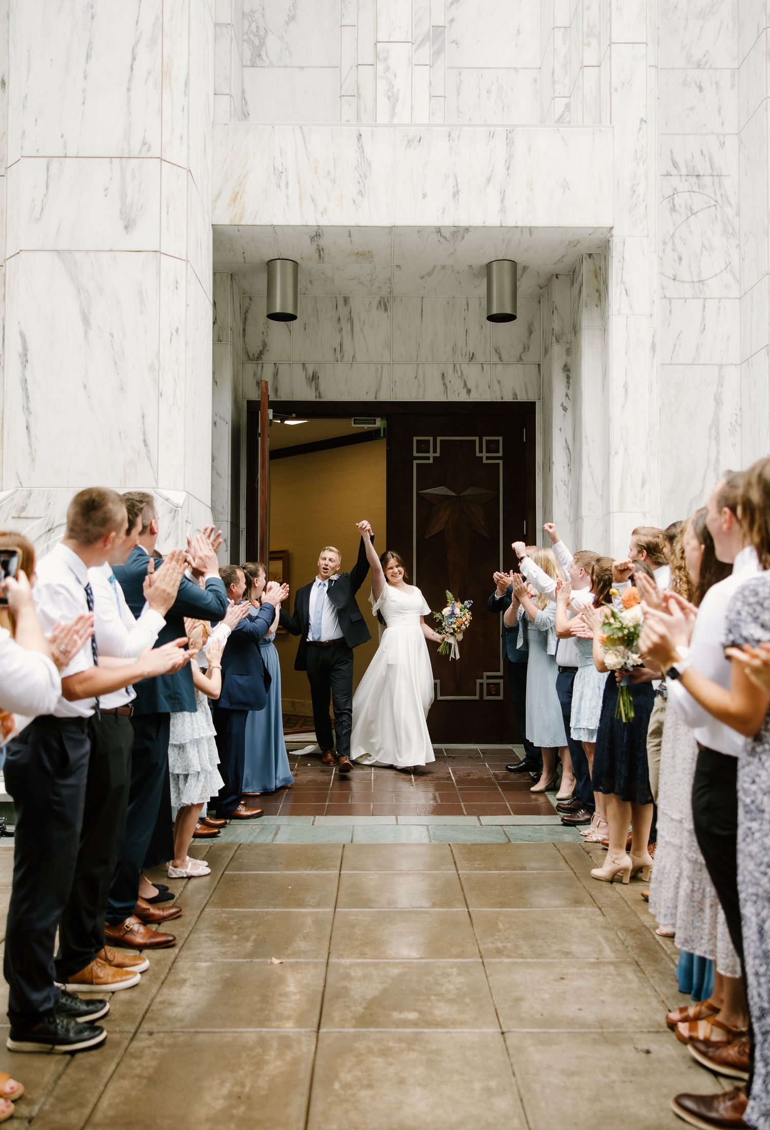 Newlyweds celebrate their wedding exit through a marble hall as guests clap and cheer.