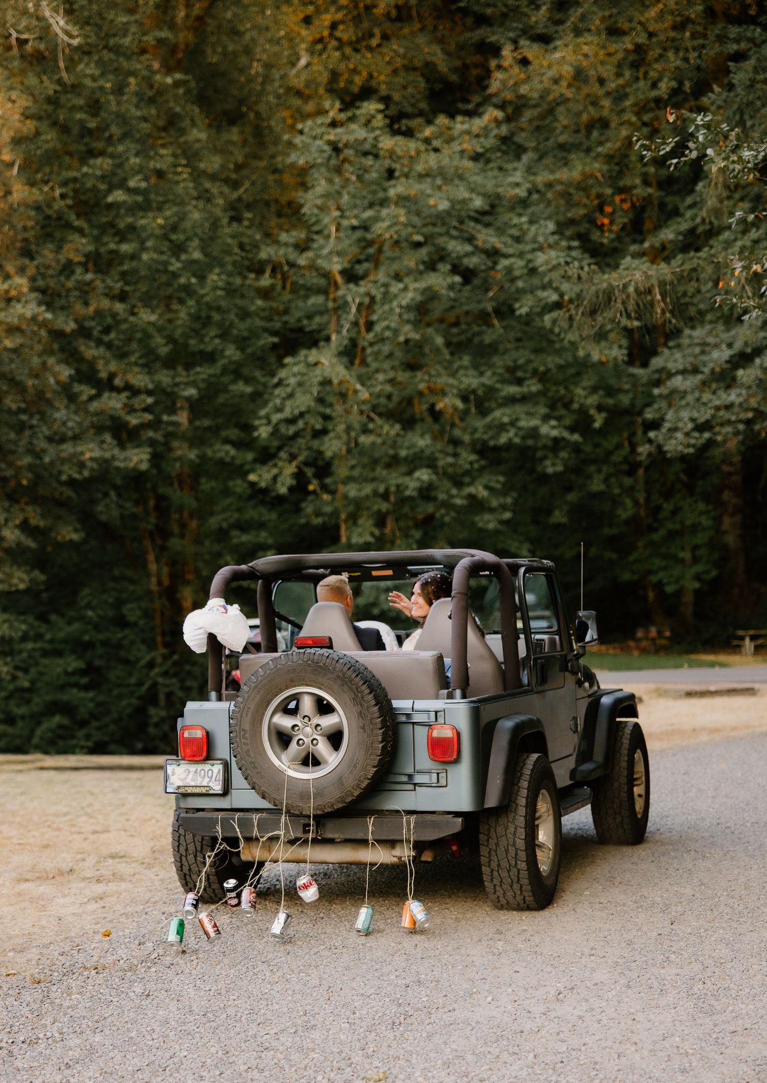 Bride and groom ride in a decorated gray Jeep Wrangler with hanging ornaments on a gravel forest path.