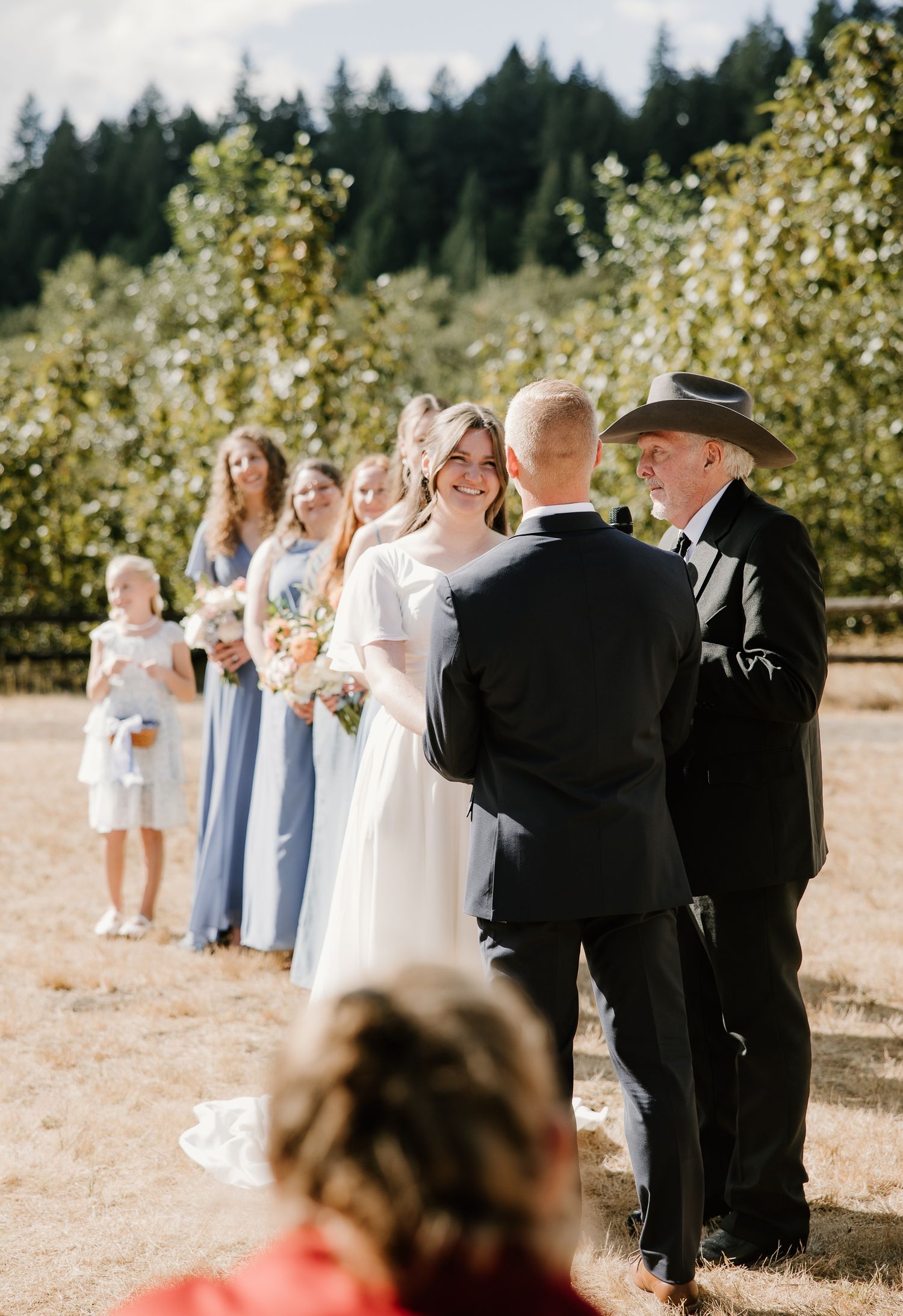 Bride and groom exchange vows outdoors with bridesmaids in blue dresses and orchard backdrop.