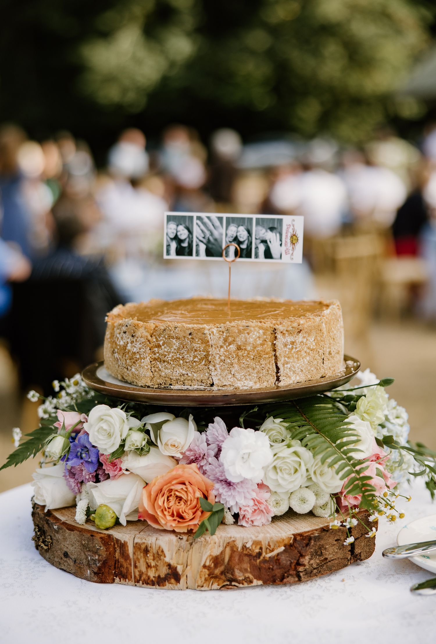Rustic wedding cheesecake topped with photos on wood slice surrounded by colorful fresh flowers.