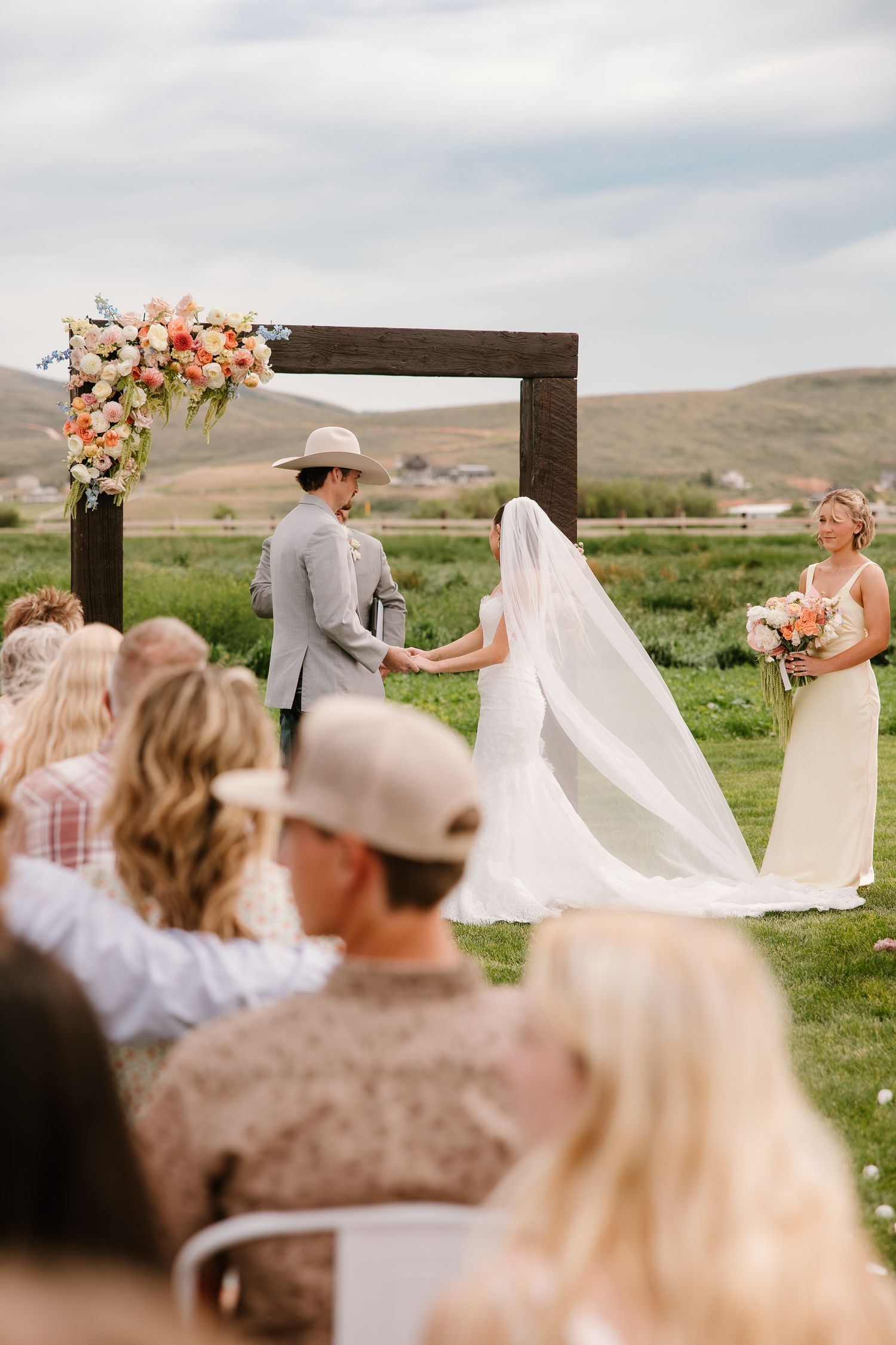 Bride and groom hold hands at wooden arch altar adorned with peach floral arrangements outdoors.