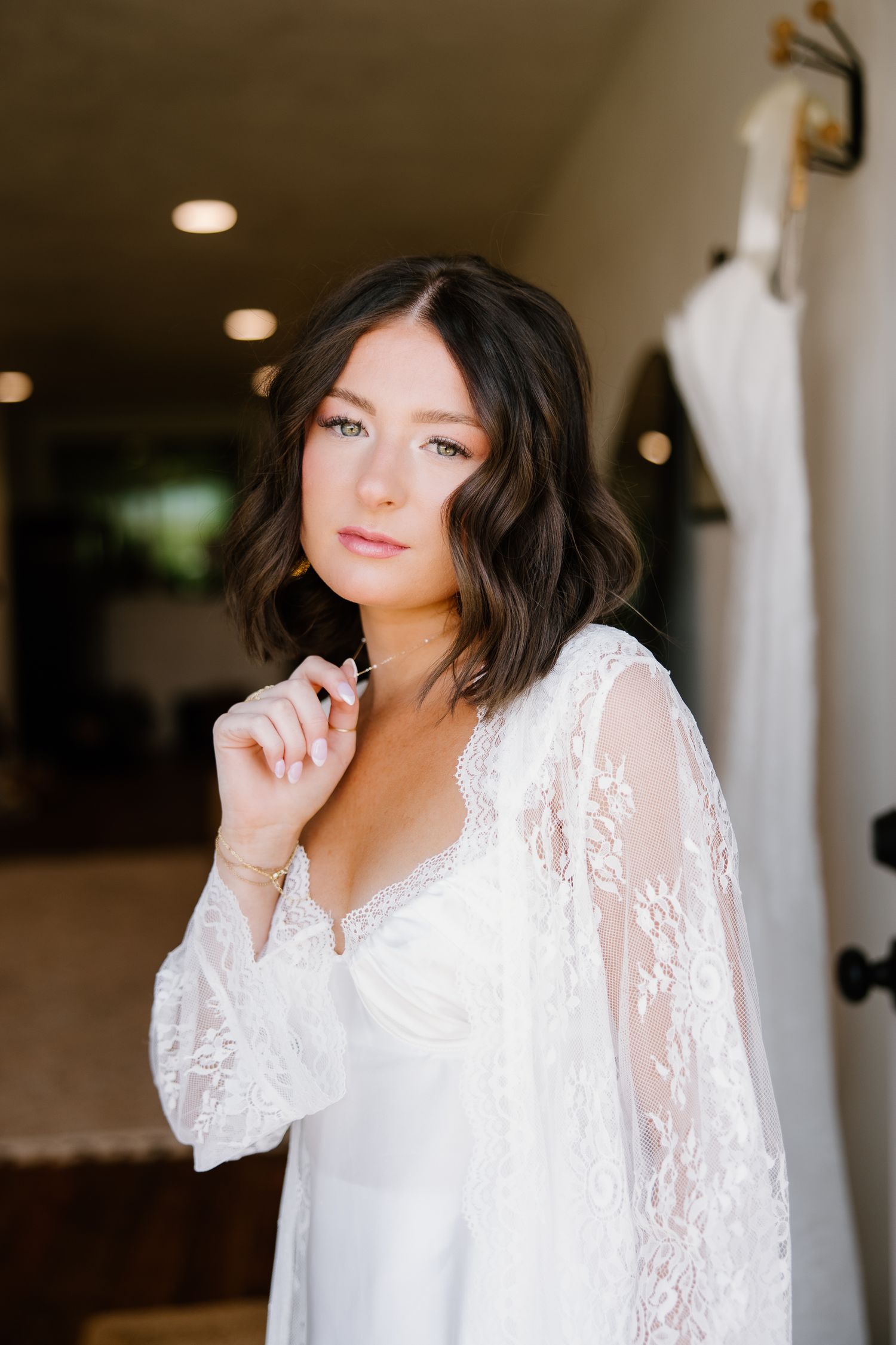 Bride in white lace robe preparing for her wedding day, posing elegantly indoors.