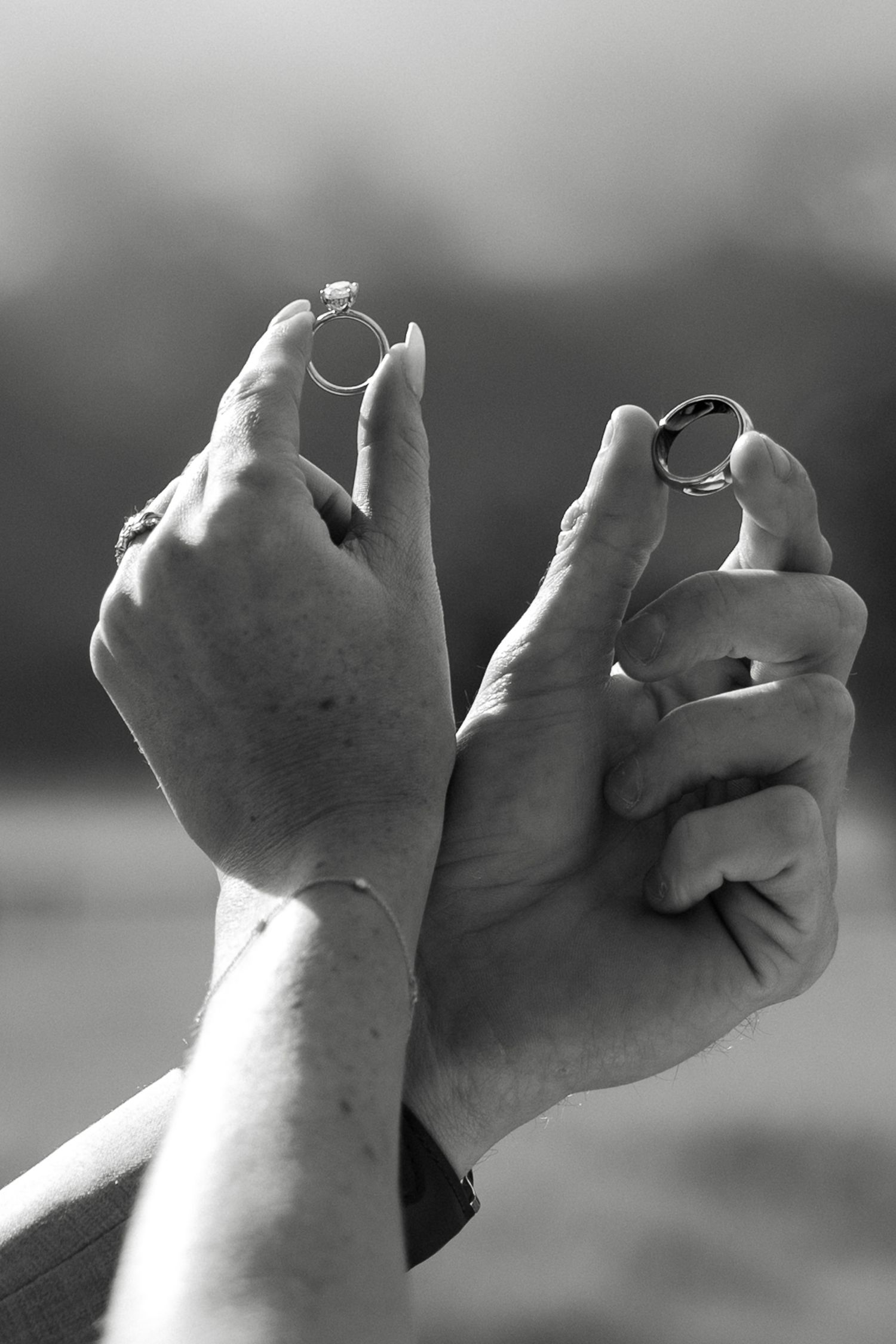 Black and white close-up of a bride and groom holding up their wedding rings against a blurred outdoor background.