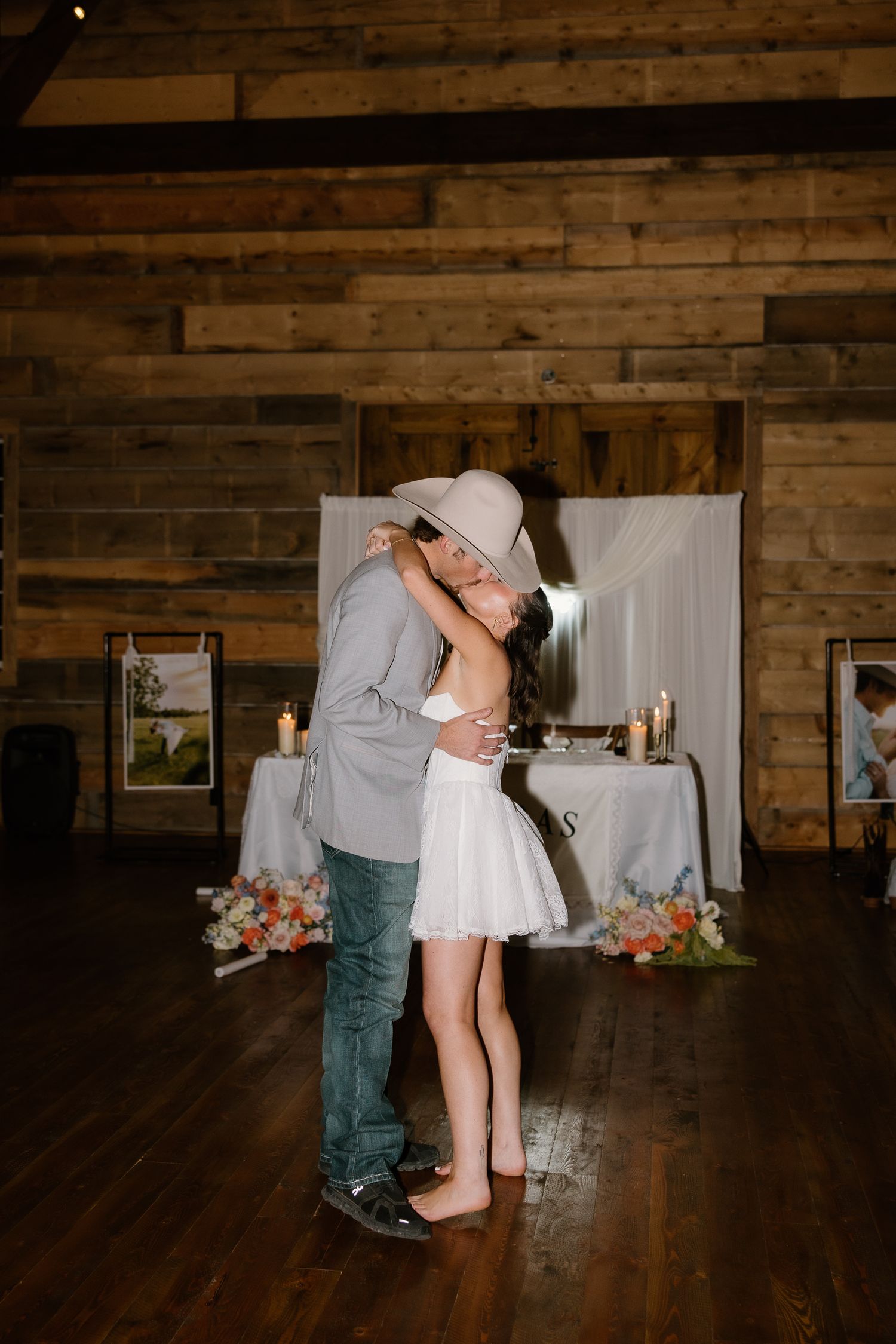 Newlyweds share first dance kiss in rustic barn venue, groom in cowboy hat and gray suit.