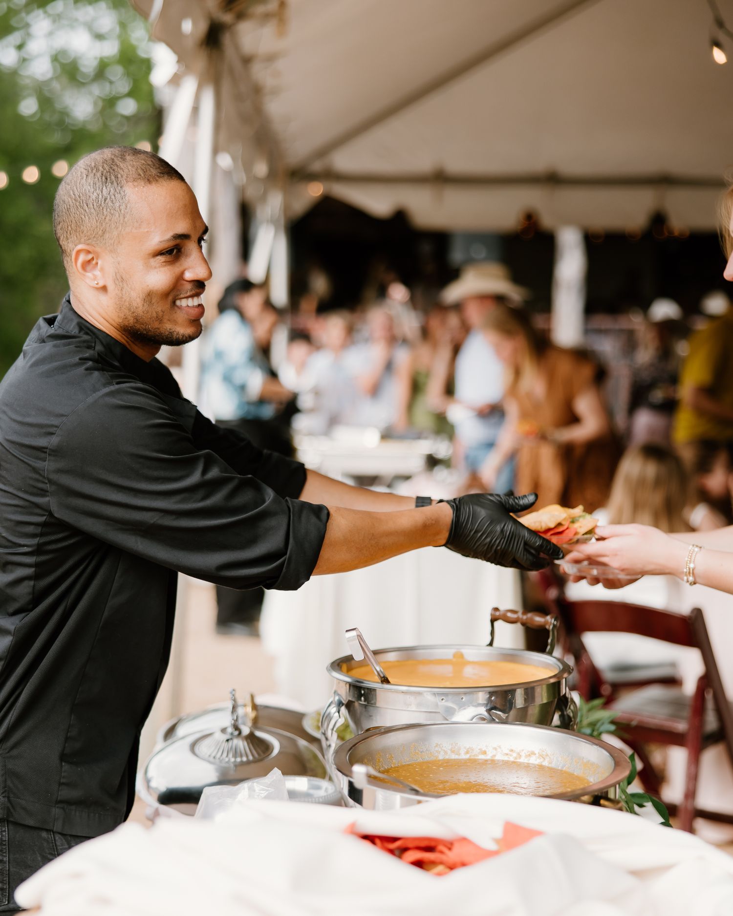 Smiling catering staff member in black uniform serves food at an elegant outdoor wedding reception.