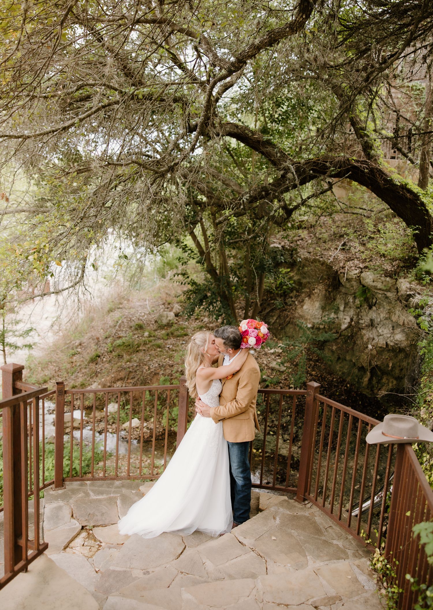 Bride and groom kissing on a stone balcony surrounded by lush trees at an outdoor wedding venue.