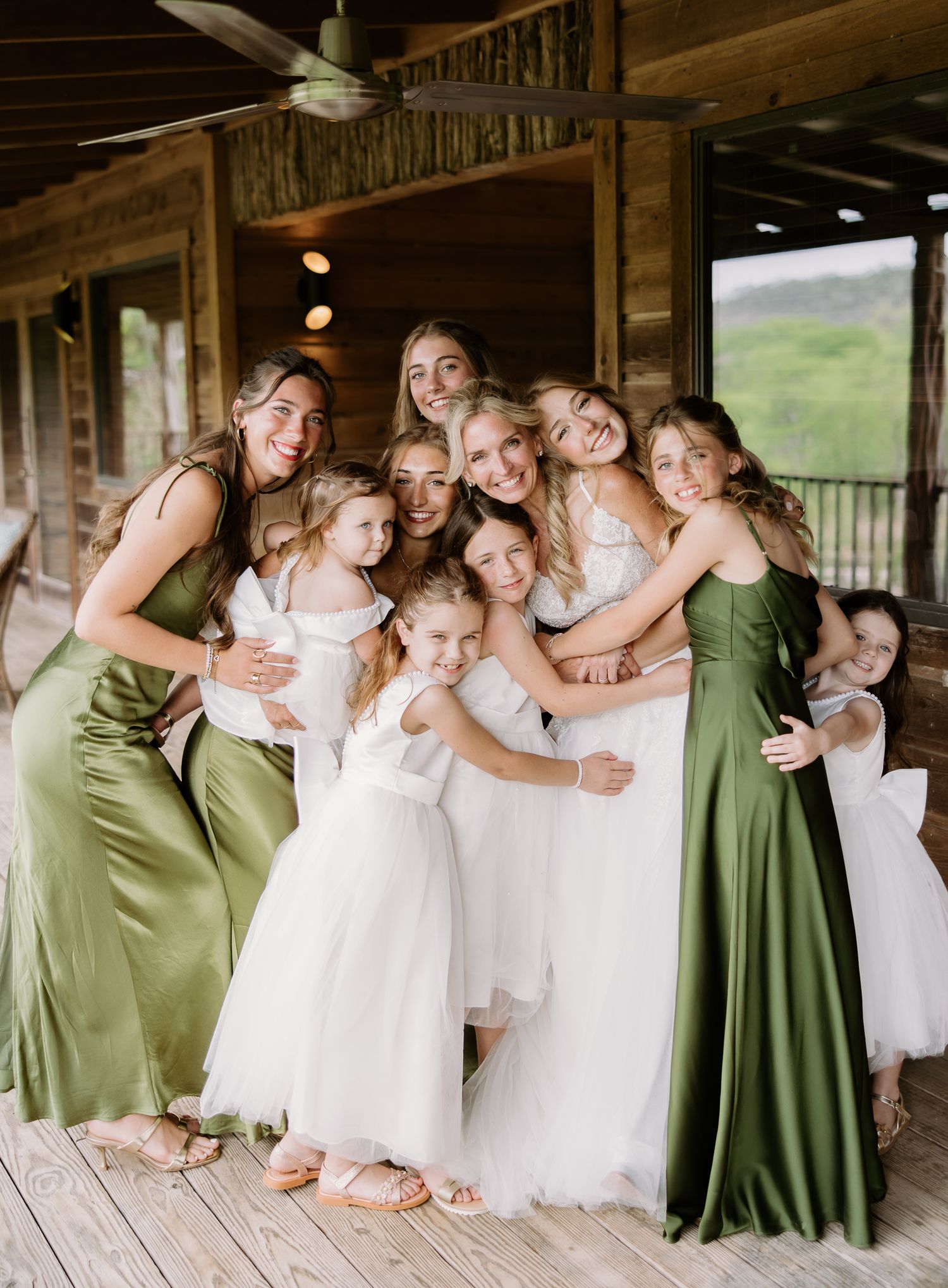 Bridesmaids in green satin dresses pose with flower girls in white on a rustic wooden porch at a wedding.