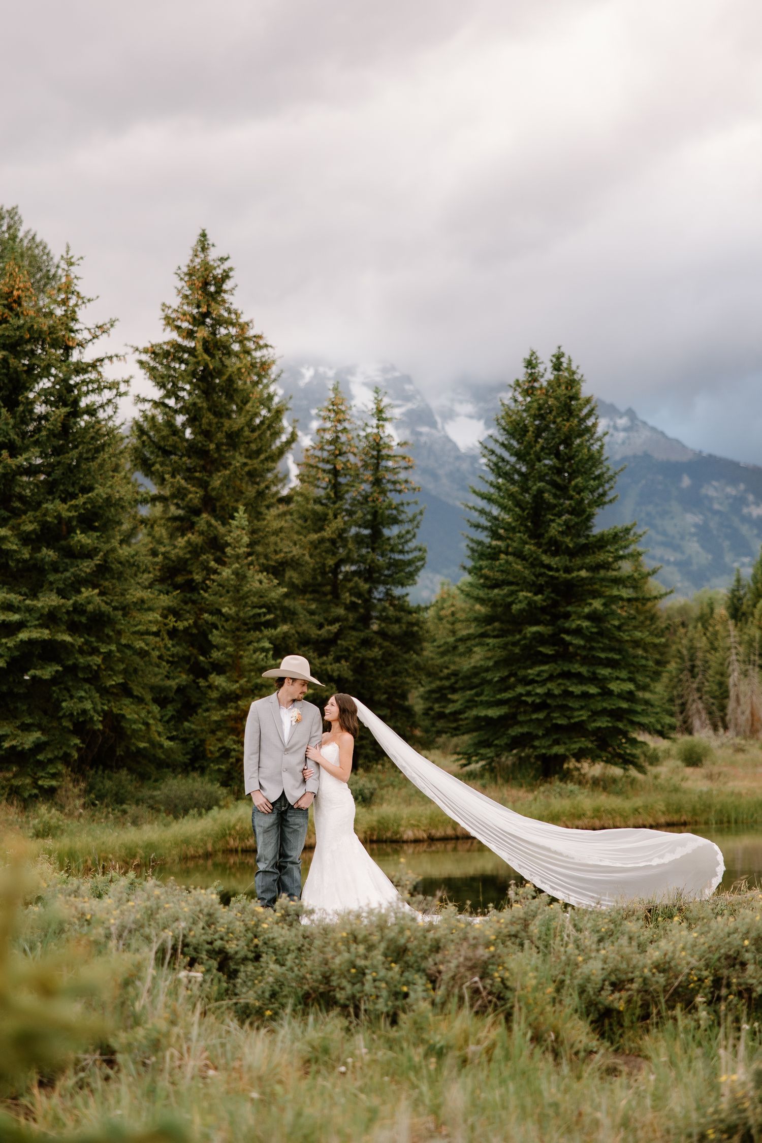 Bride and groom stand in a meadow with veil flowing in wind, evergreen trees and misty Grand Teton mountains behind them.