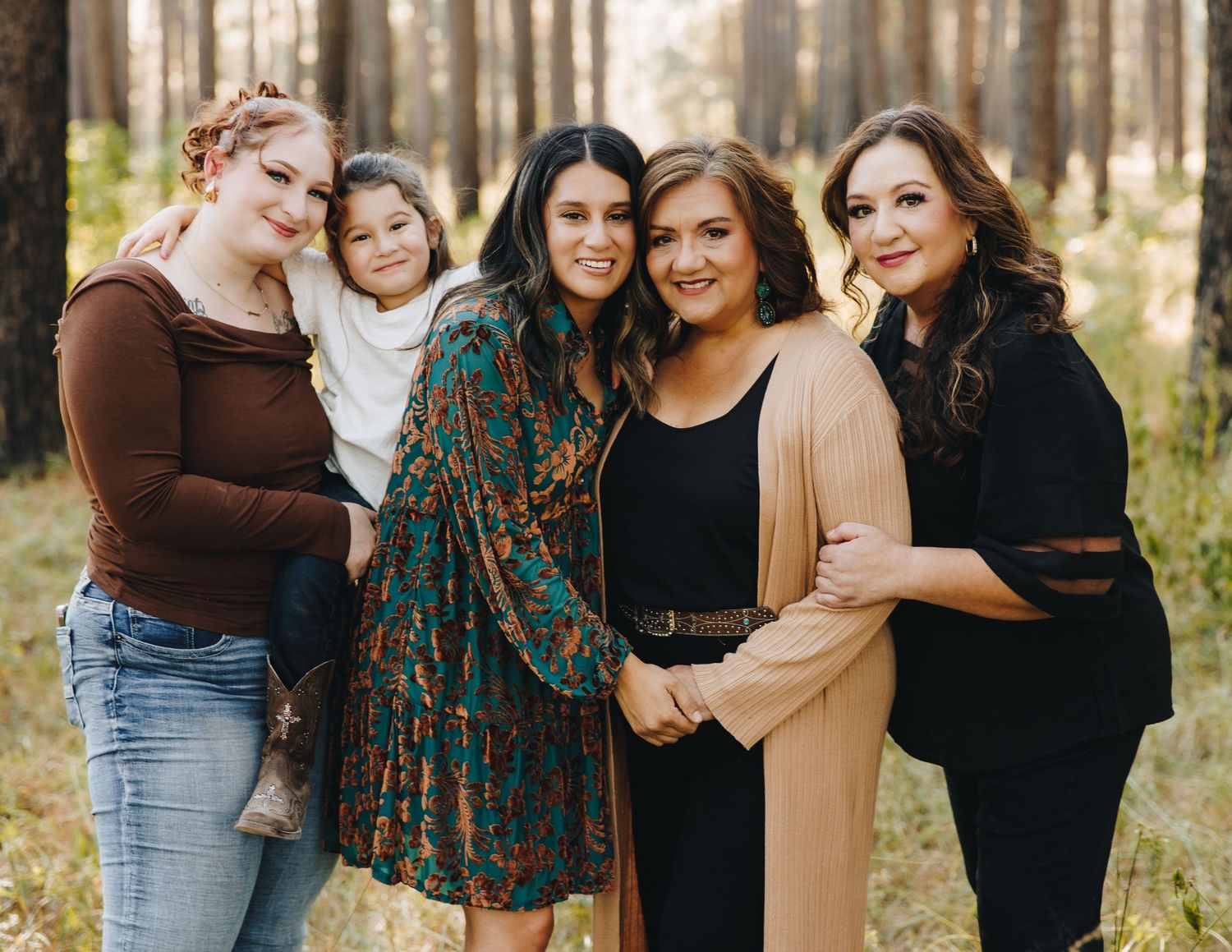 Five women of different ages posing together smiling in a pine forest setting.