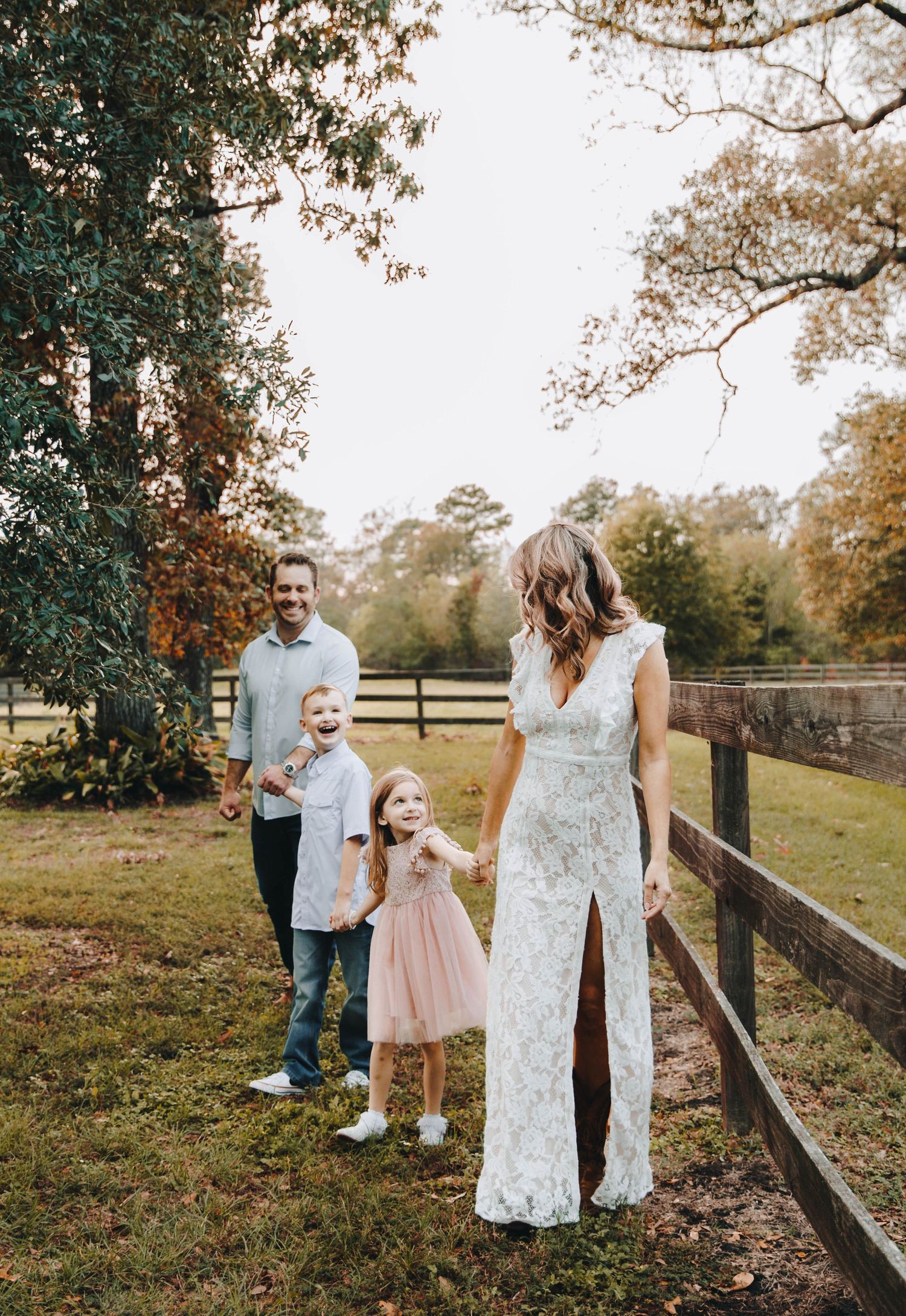 Family of four walking along wooden fence in autumn, mother in white lace dress, children smiling.