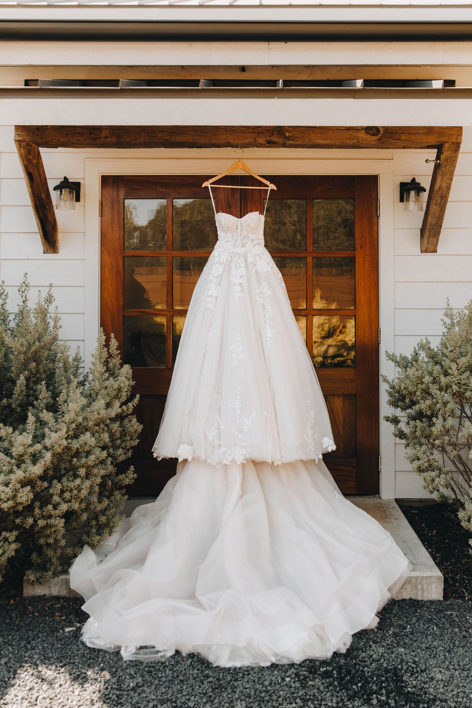 Elegant strapless lace ballgown wedding dress hanging on wooden door of a white farmhouse building.