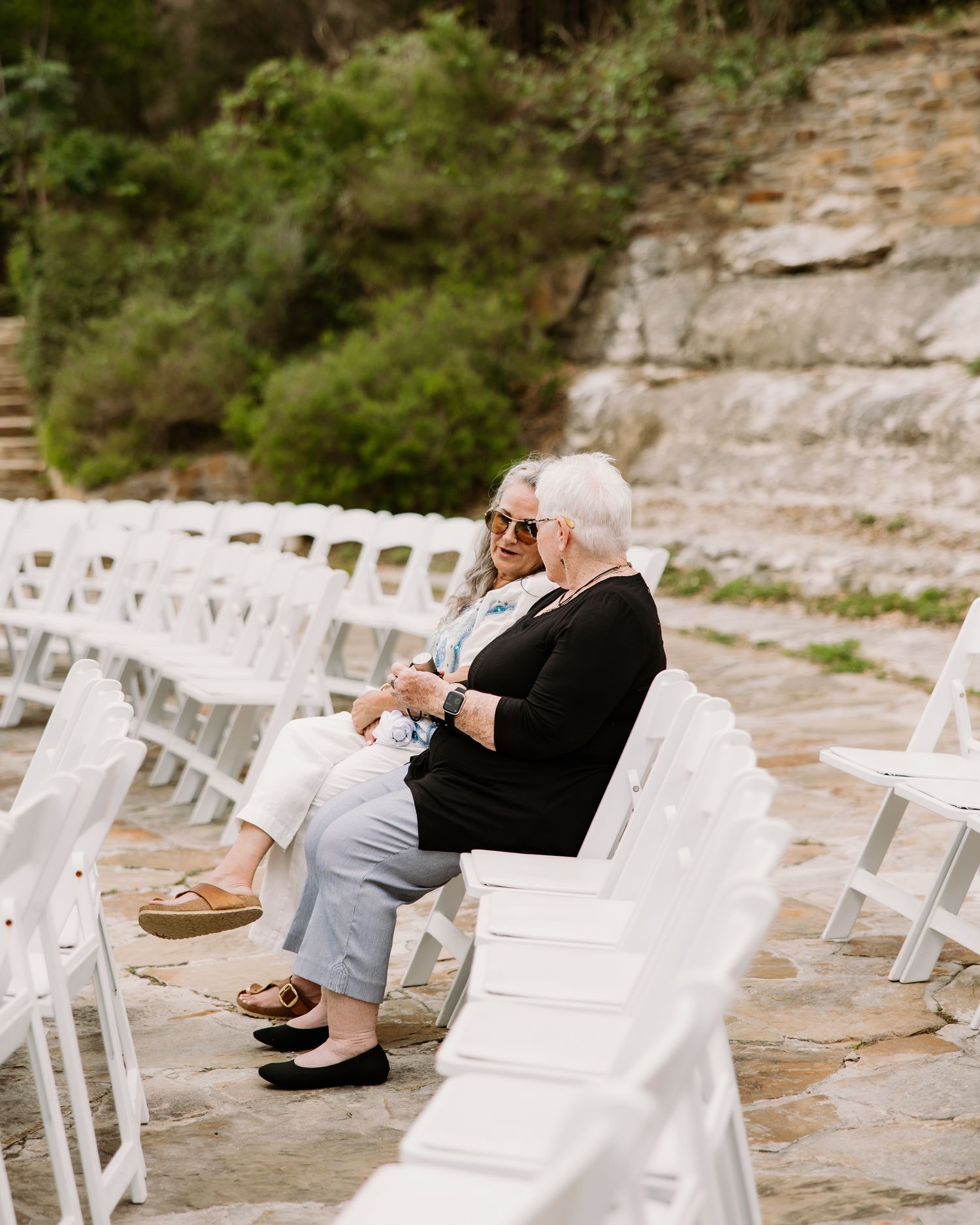 Two elderly women sit together in white chairs at an outdoor wedding ceremony venue with stone steps.