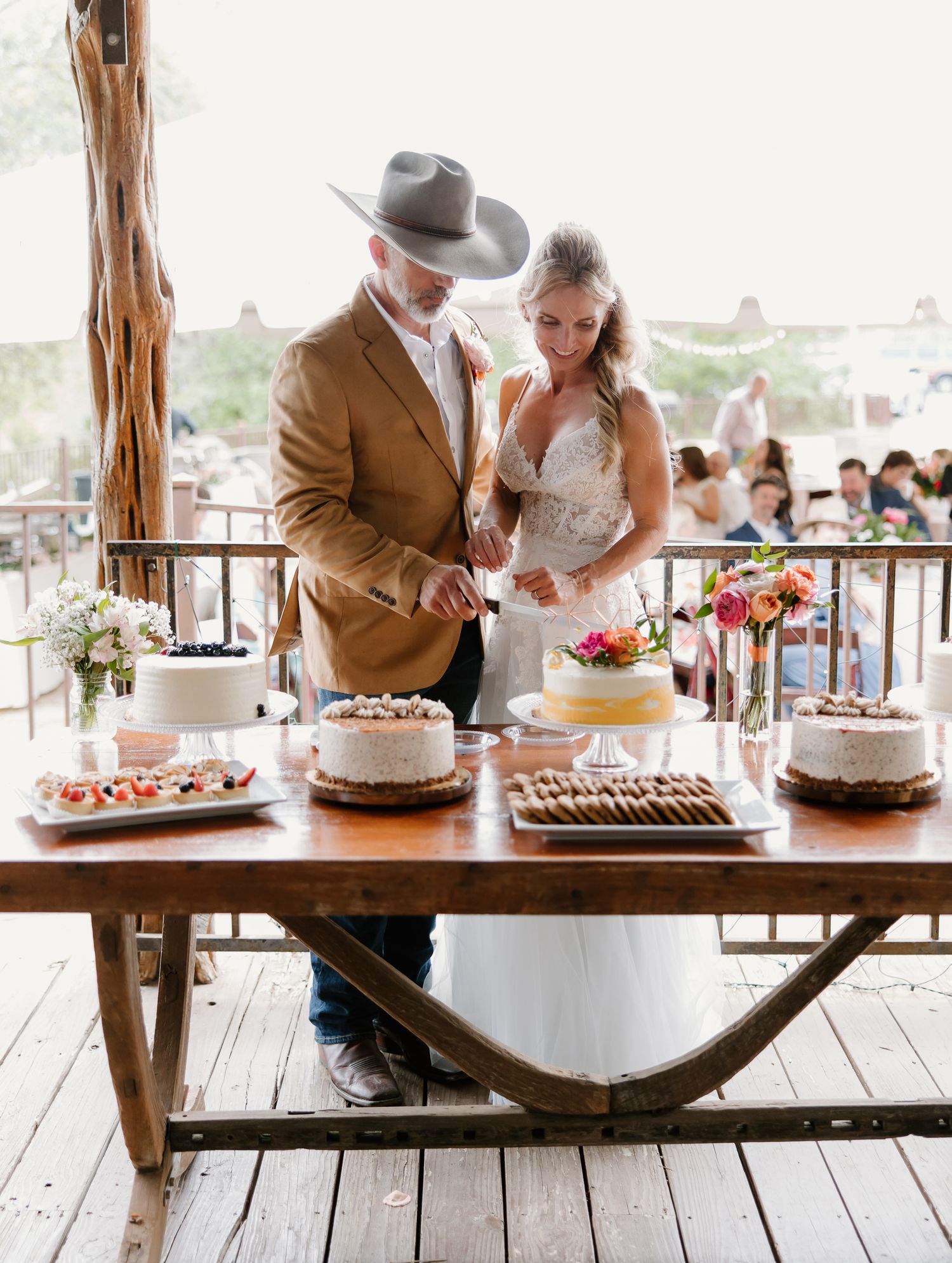 Bride and groom cutting wedding cake at a rustic wooden dessert table with multiple cakes under an open-air pavilion.