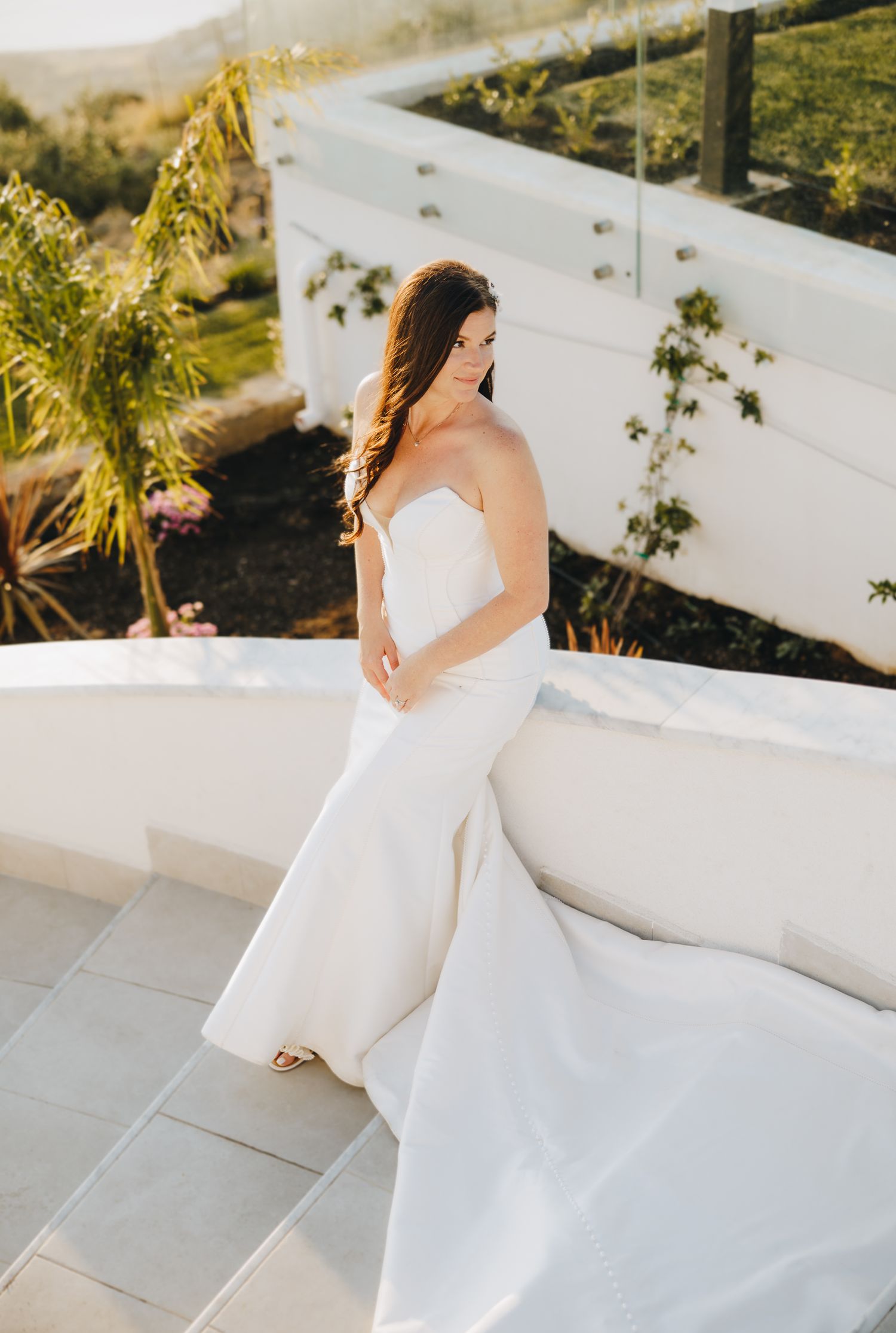 Bride in strapless white mermaid wedding gown posing on sunlit terrace with lush greenery backdrop.