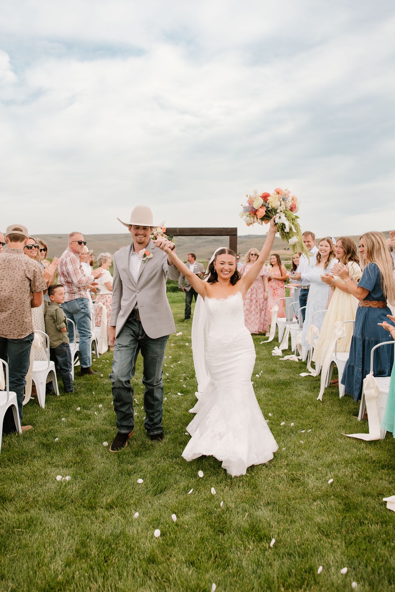 Newlyweds celebrate recessional with raised hands and bouquet on a green lawn at an outdoor wedding.